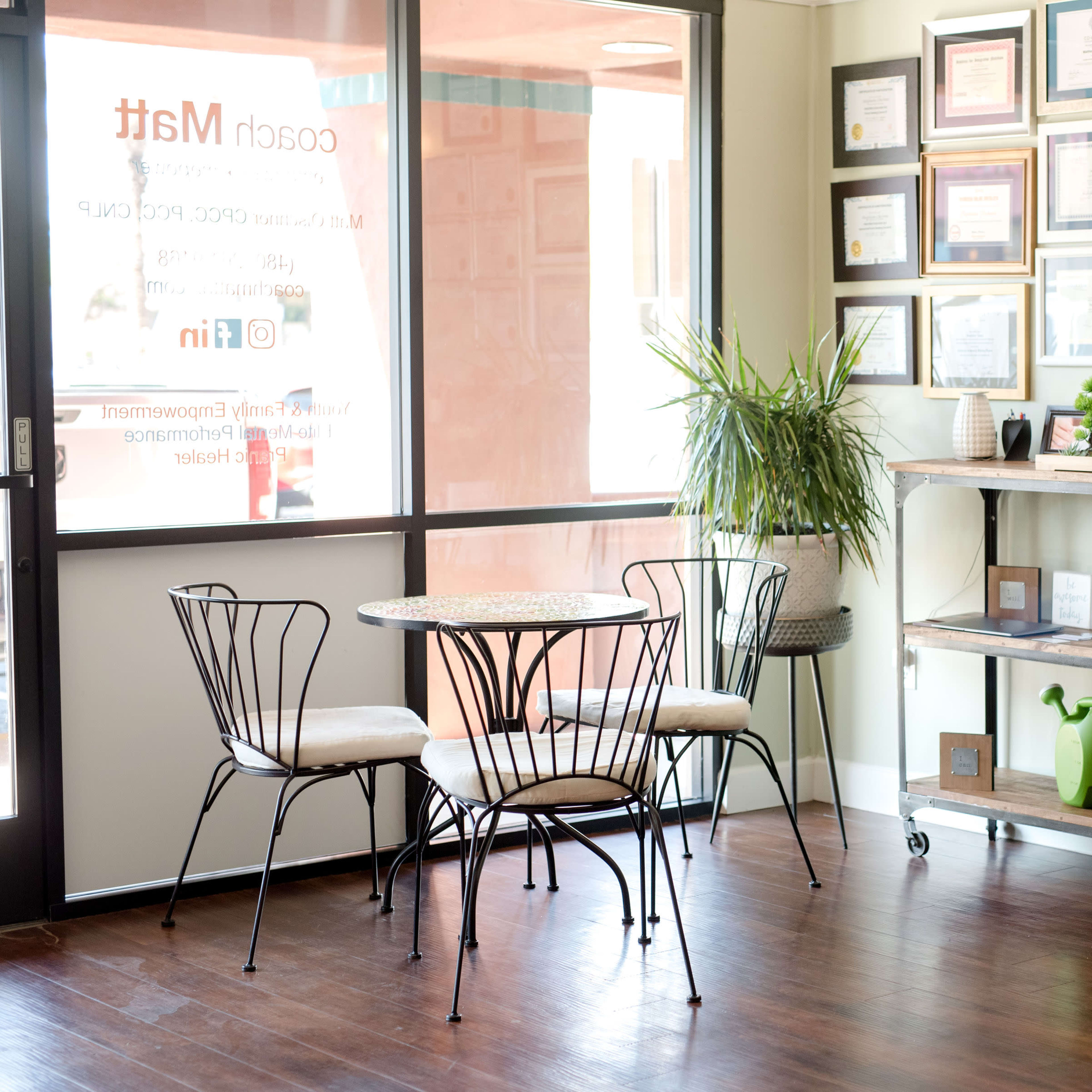 A small waiting area features a round table with four chairs, a plant stand, and a wall displaying various framed certificates.