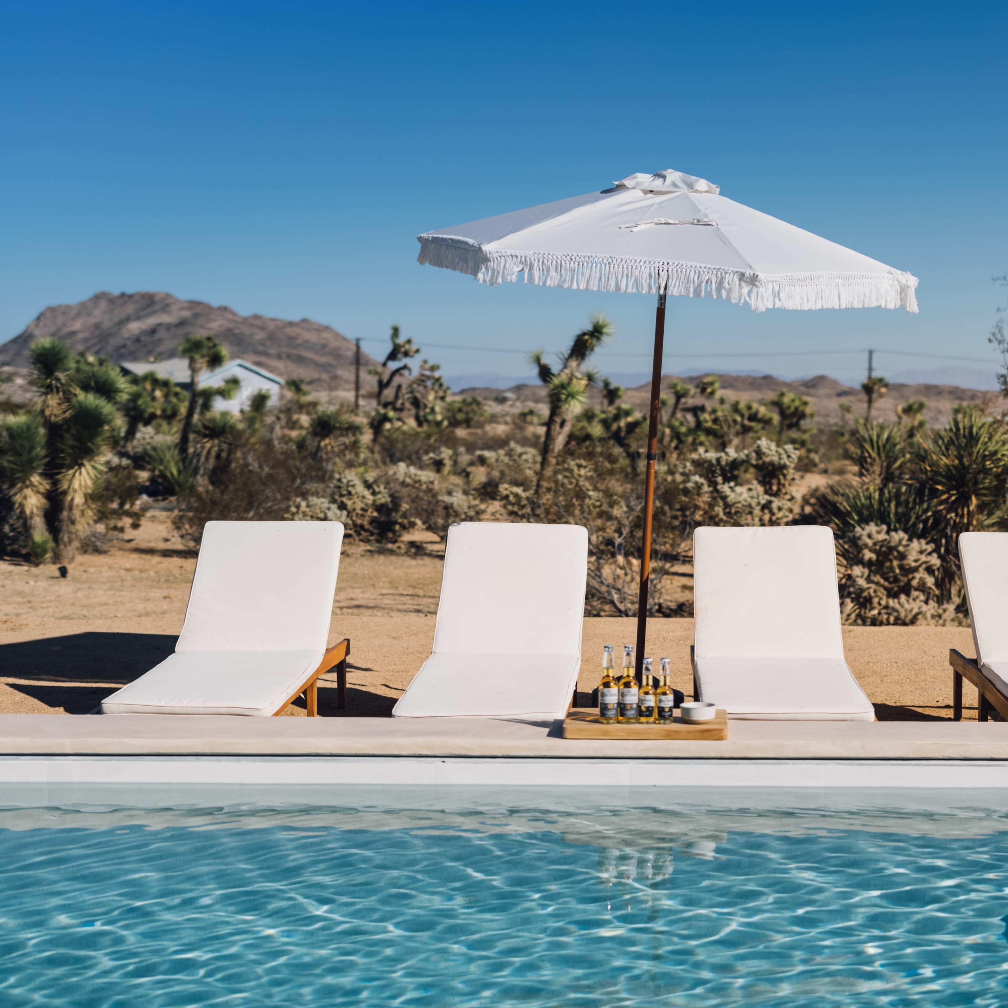 Four lounge chairs beside a swimming pool, shaded by a white umbrella, with a desert landscape and mountains in the background.