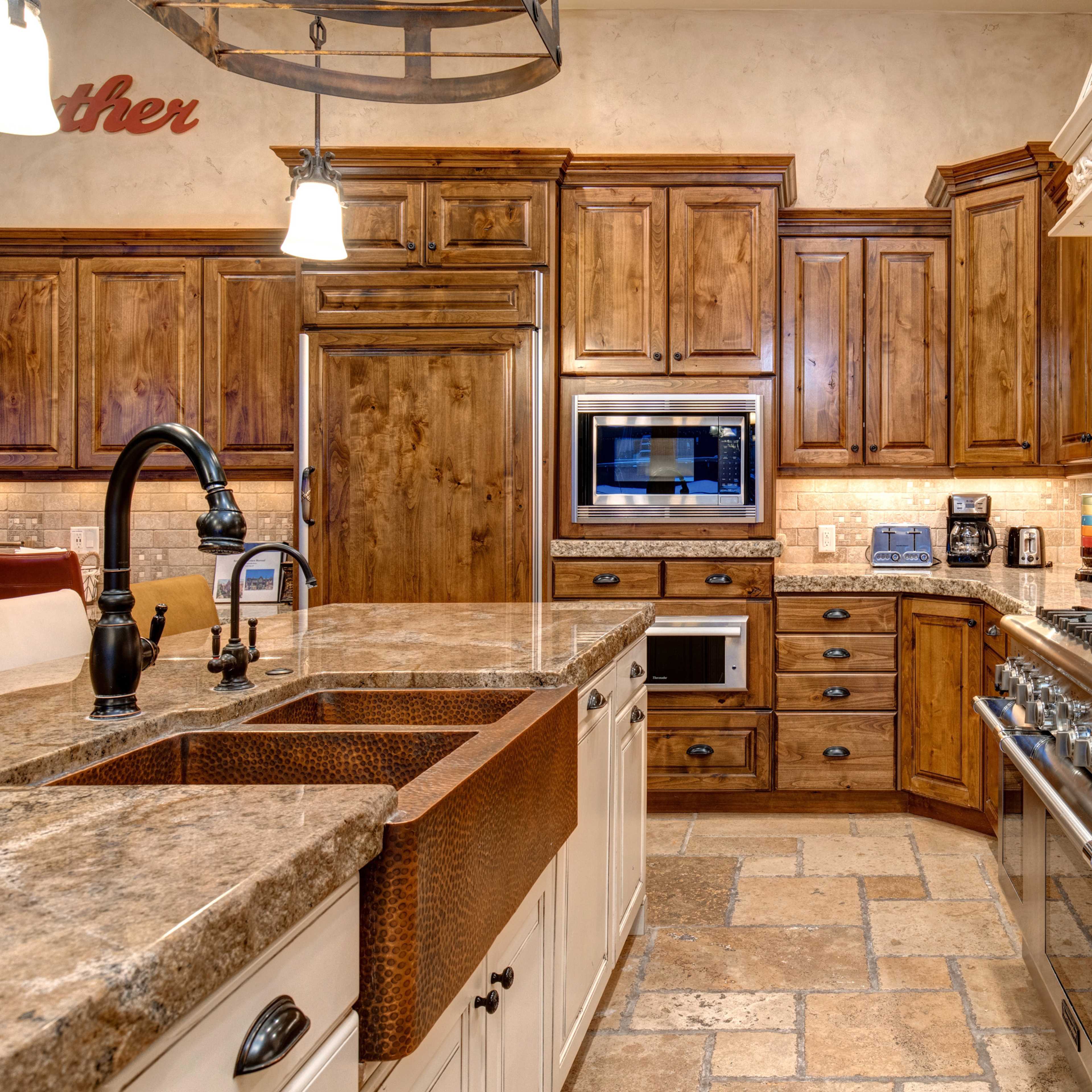 A kitchen with wooden cabinets, a large stainless steel stove, a double-basin copper sink, and stone tile flooring.