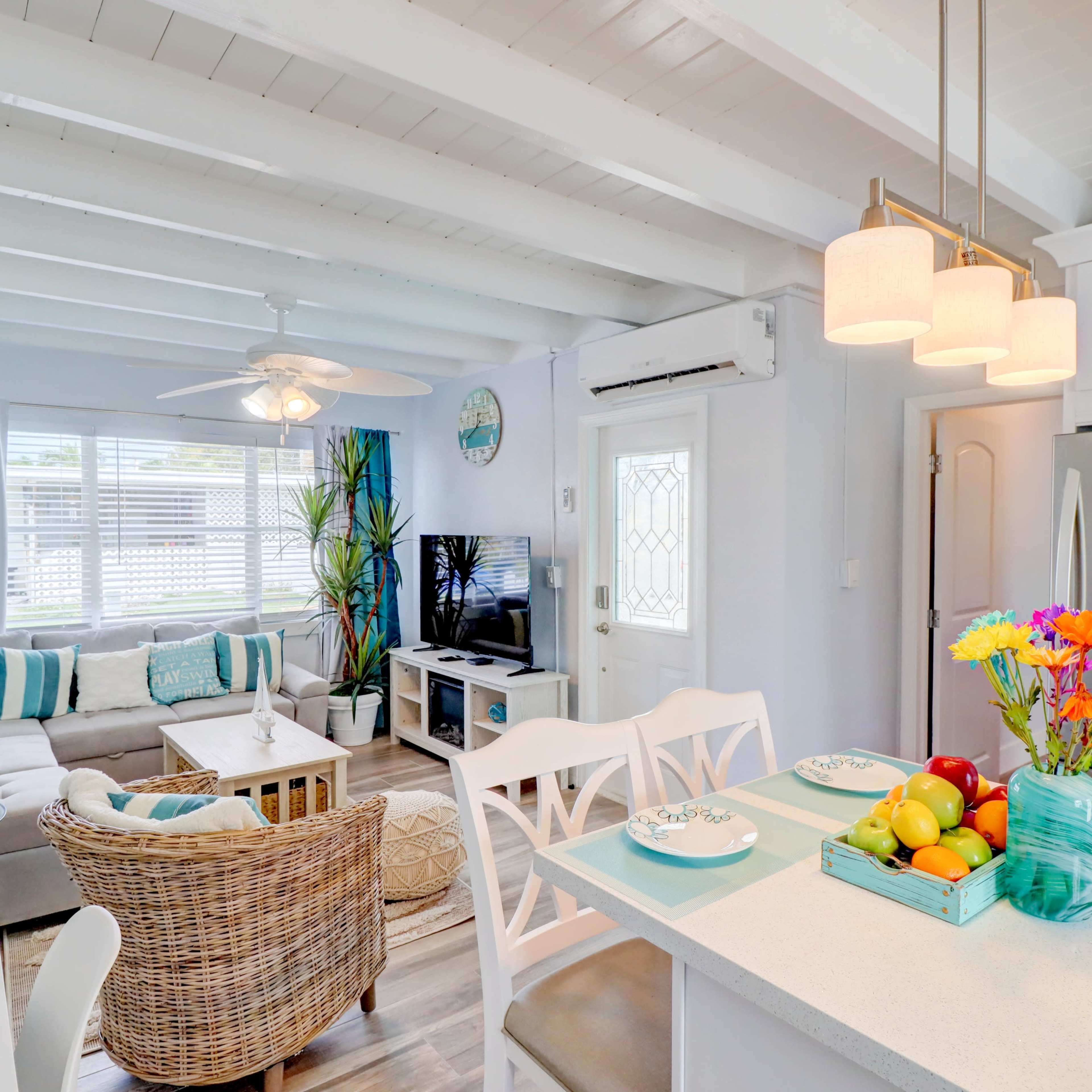 A bright, modern living room with a cozy sofa, a dining area featuring a table with fruit, and large windows letting in natural light.