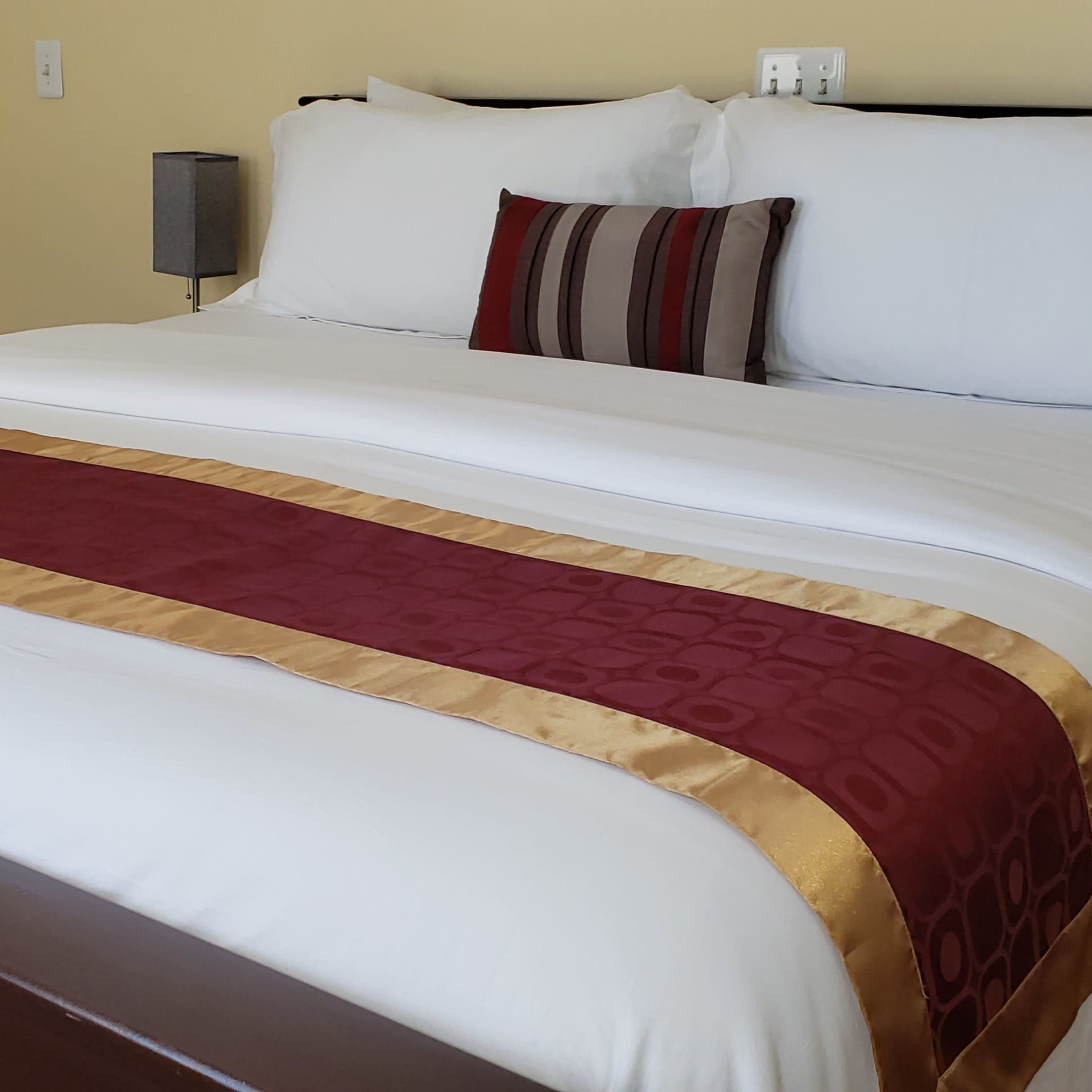 A neatly made bed with a white duvet and a burgundy and gold decorative stripe running across it, set in a softly lit bedroom.