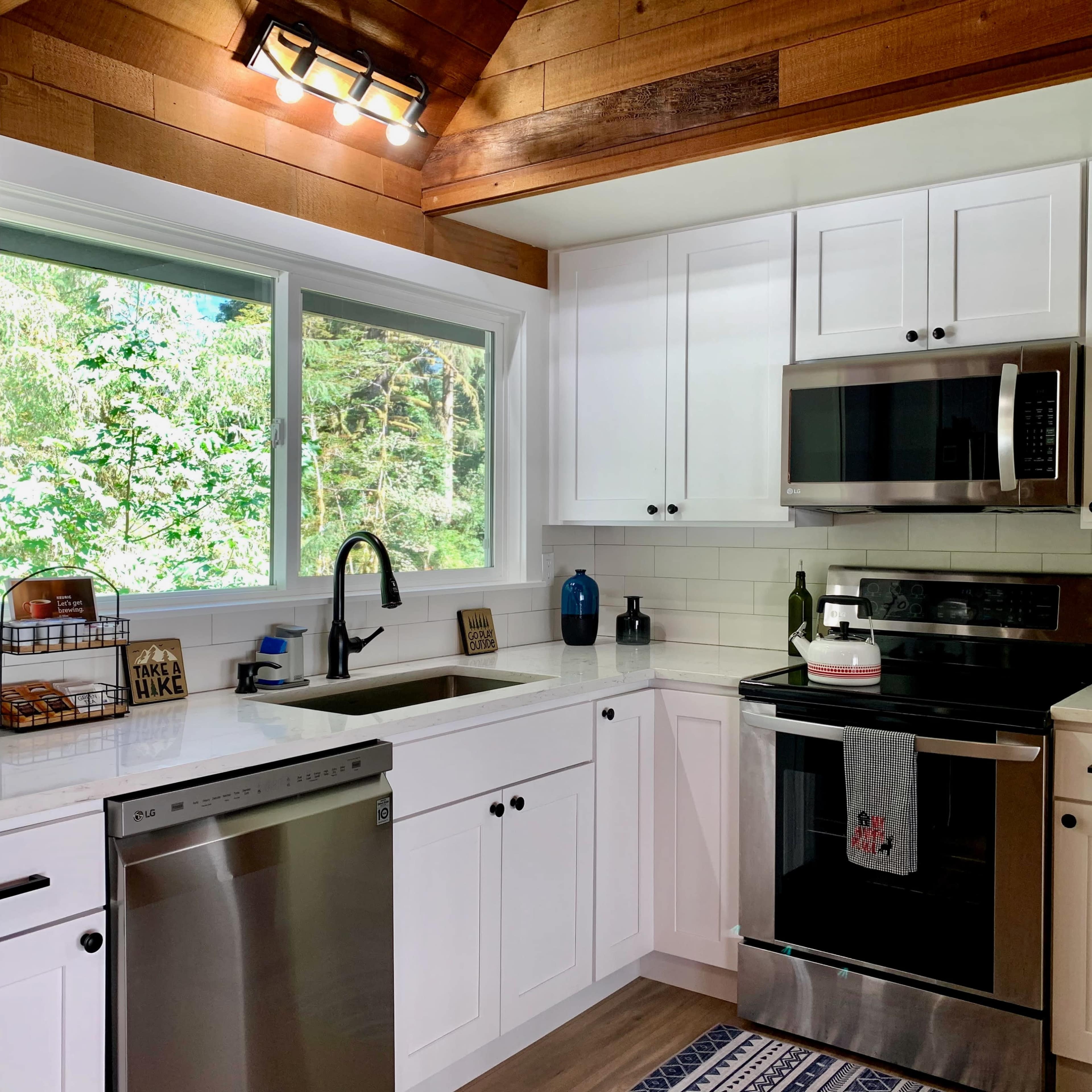 A modern kitchen with white cabinets, a stainless steel refrigerator and oven, and a window overlooking trees.