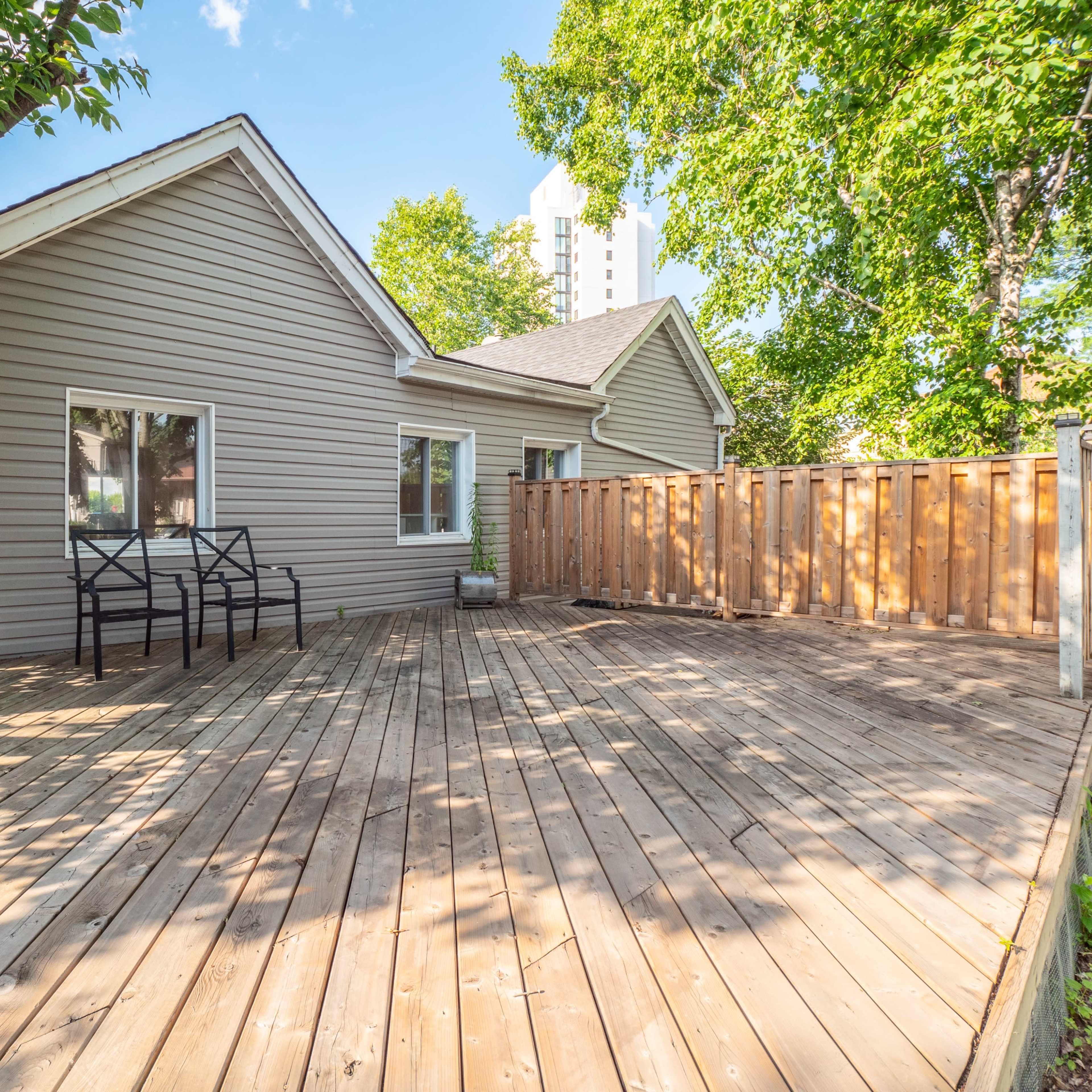 A wooden deck area next to a single-story house, surrounded by greenery and featuring two chairs.