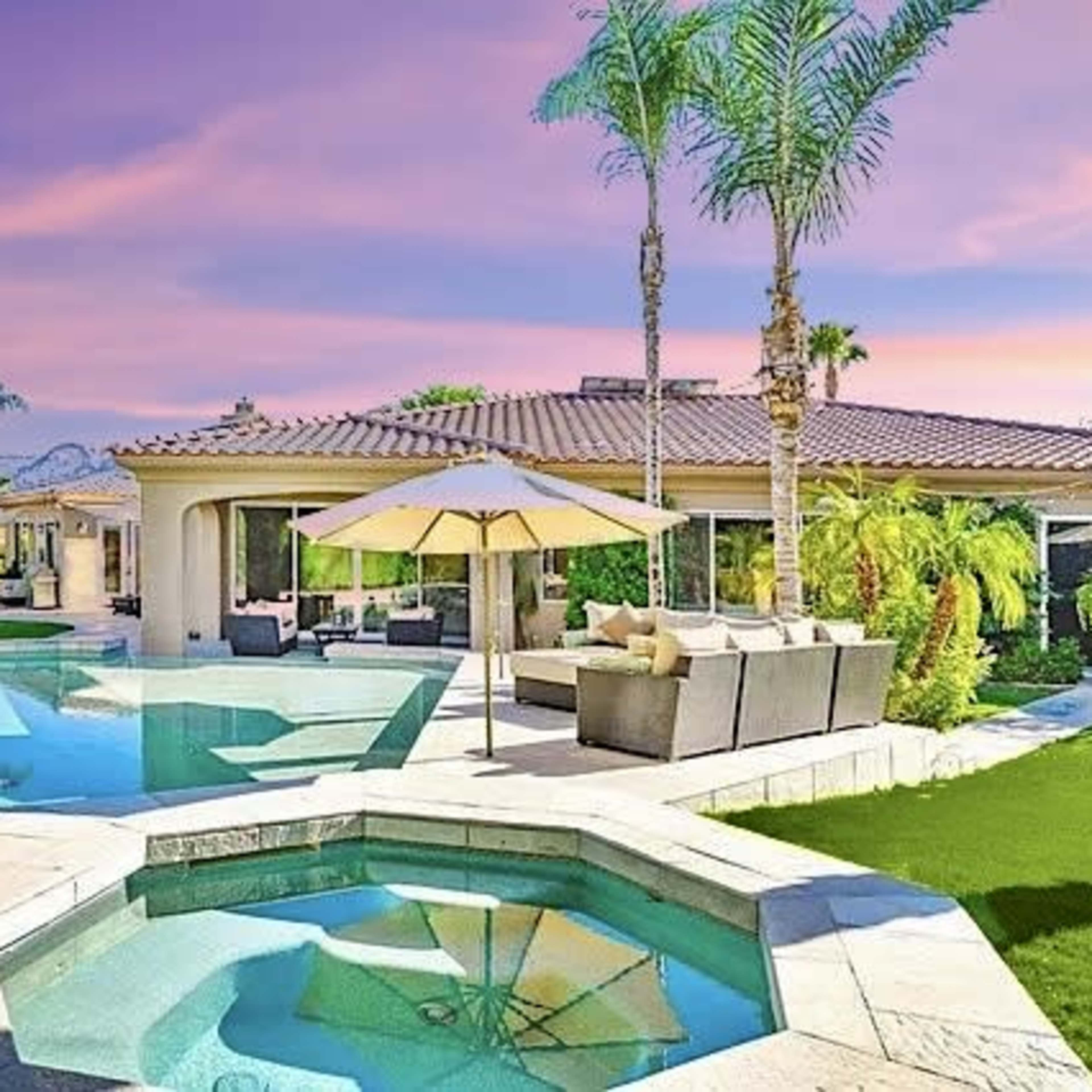 A backyard with a swimming pool, spa, and a patio area surrounded by palm trees and landscaped greenery.