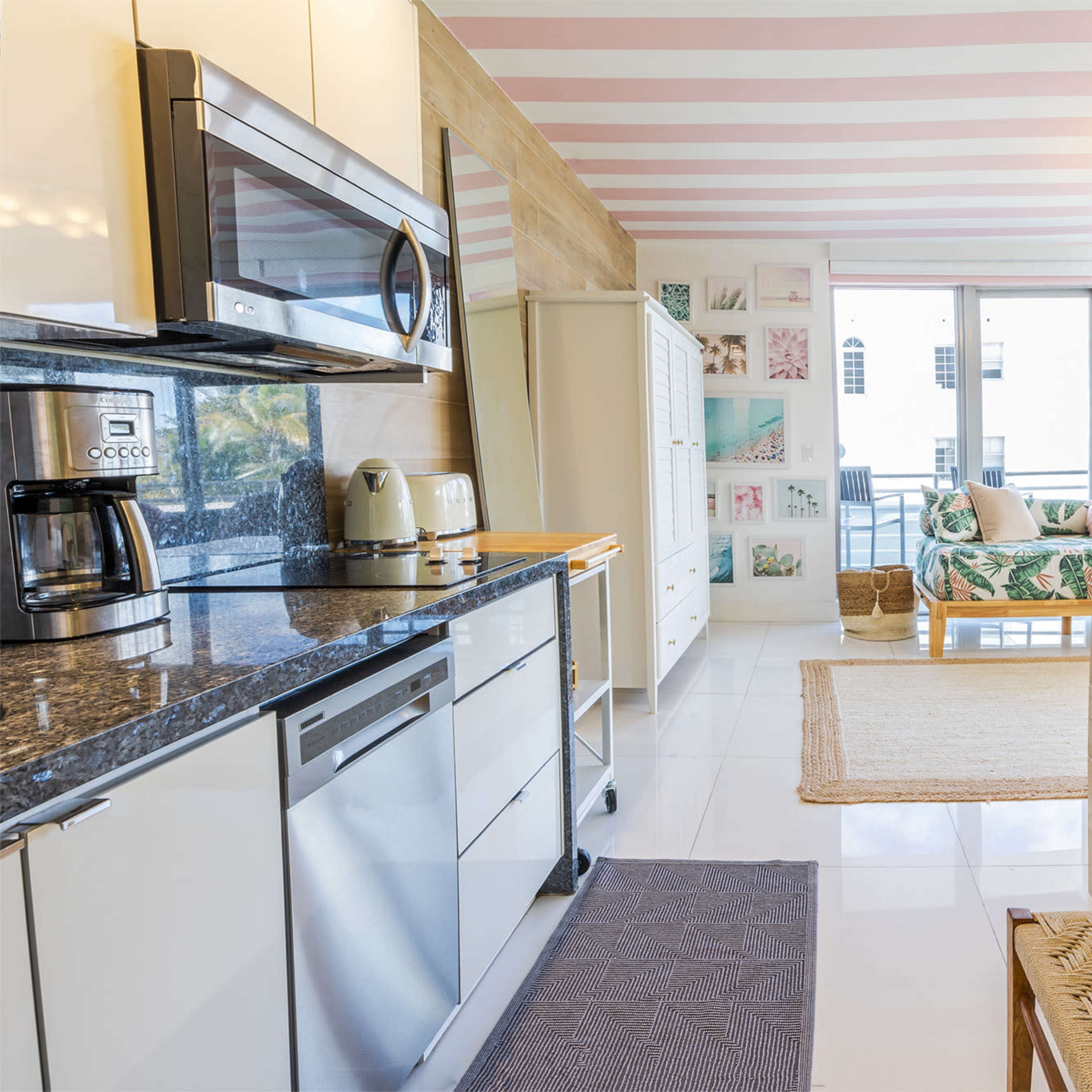 A modern kitchen with stainless steel appliances opens into a bright living area featuring a sofa and colorful wall art.