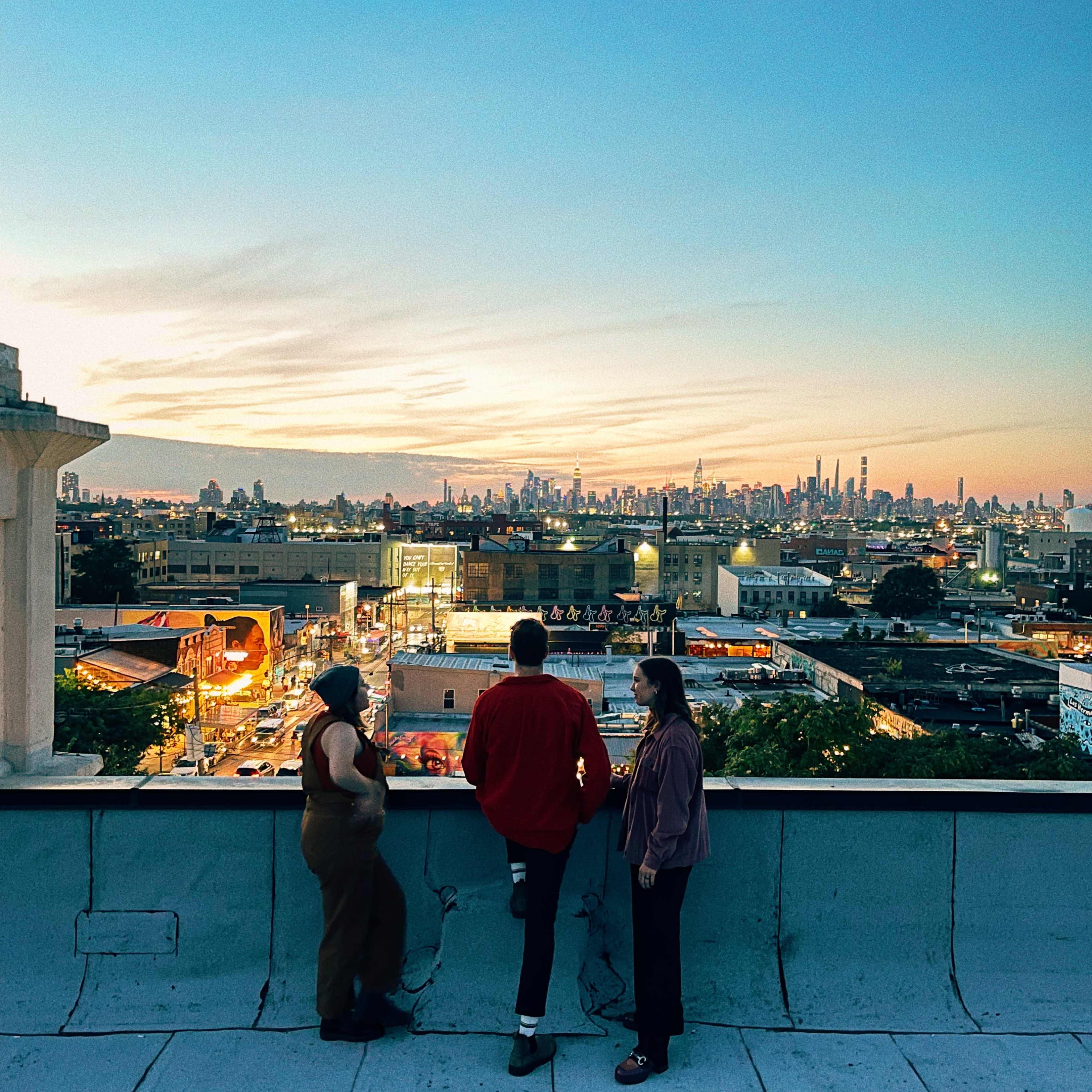 Three people stand on a rooftop looking out over a city skyline at sunset, with lights beginning to illuminate the streets below.