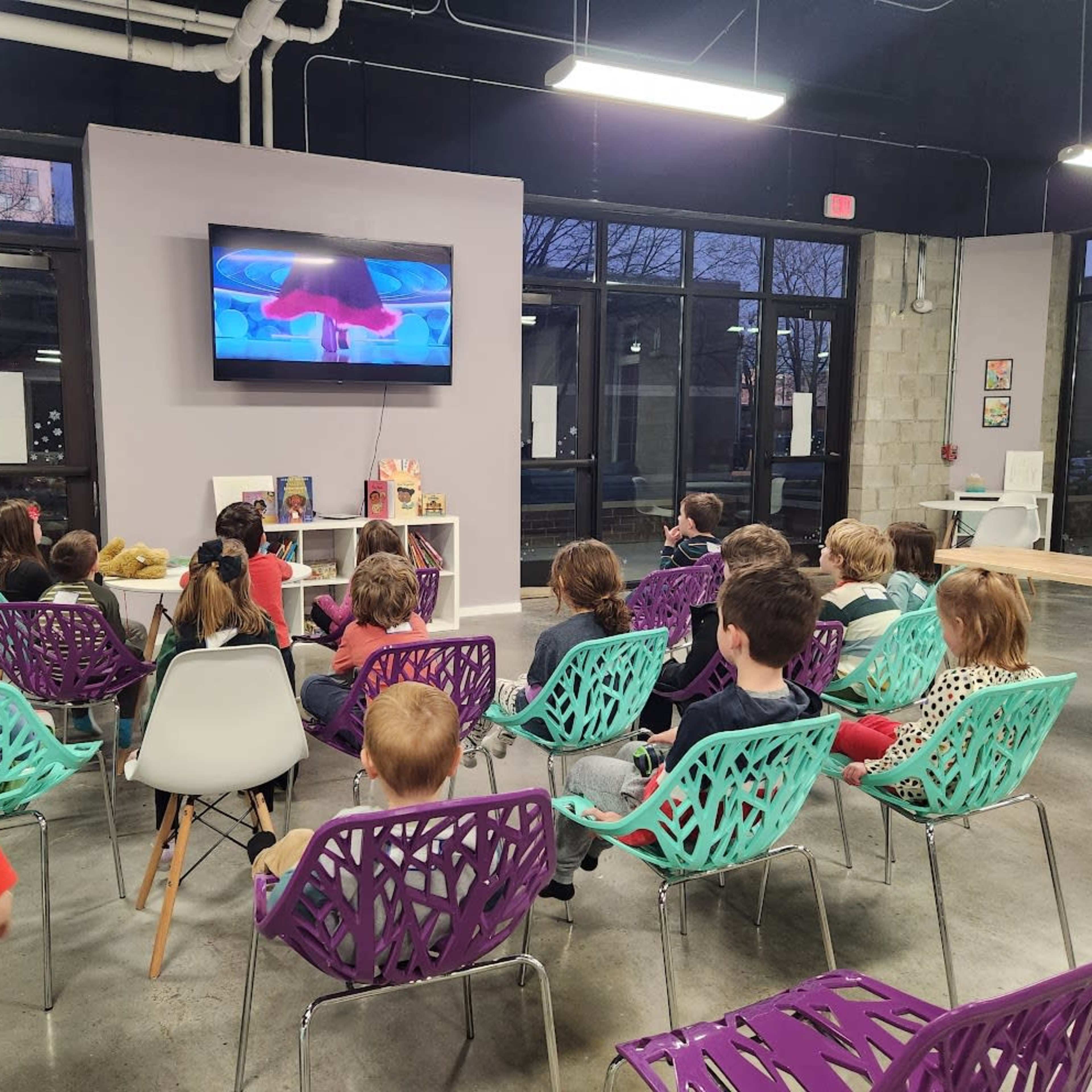 A group of children sits in colorful chairs, watching a video on a wall-mounted screen in a well-lit, modern room.
