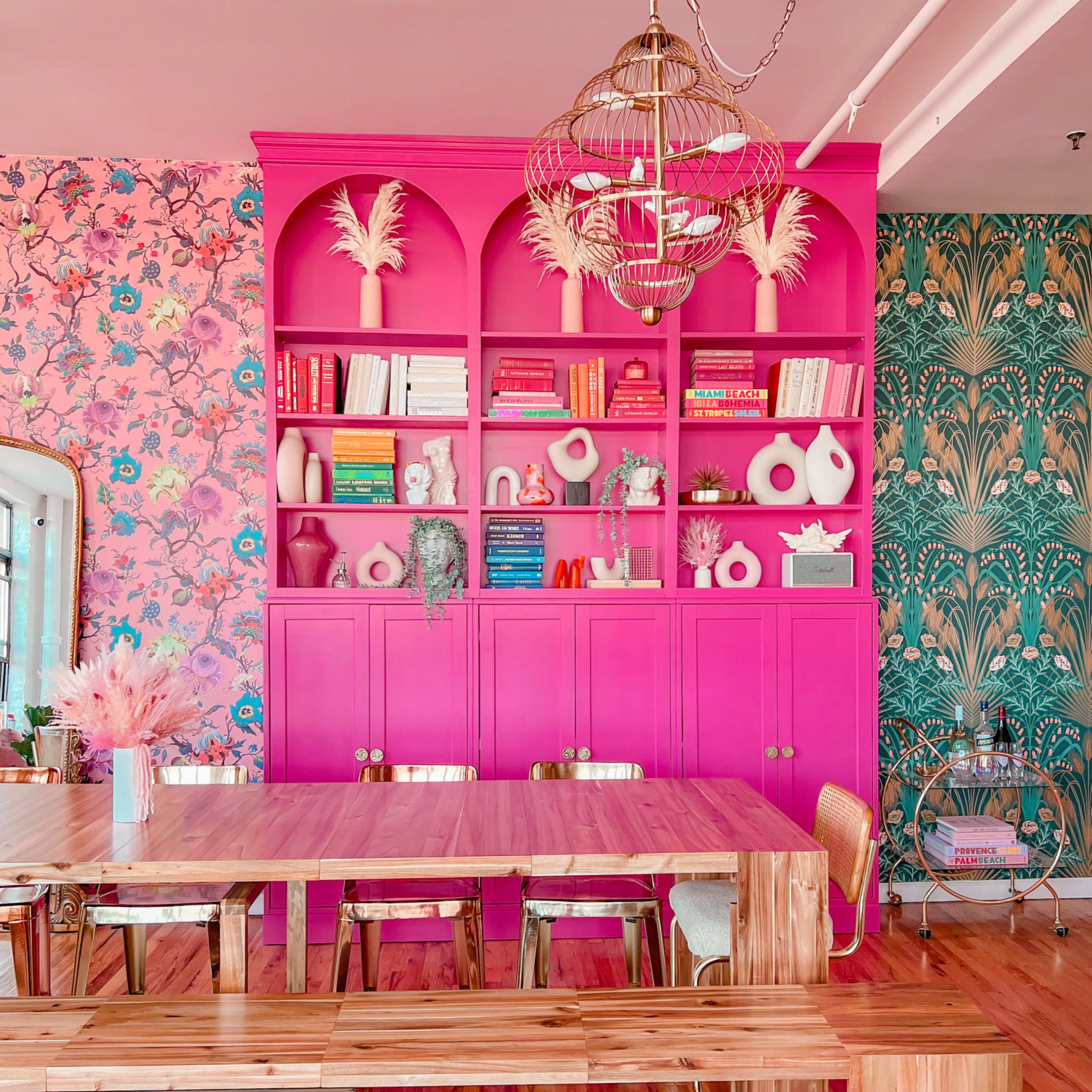 A brightly colored dining area features a pink bookshelf filled with decorative items and books, complemented by floral wallpaper and a wooden dining table with bench seating.