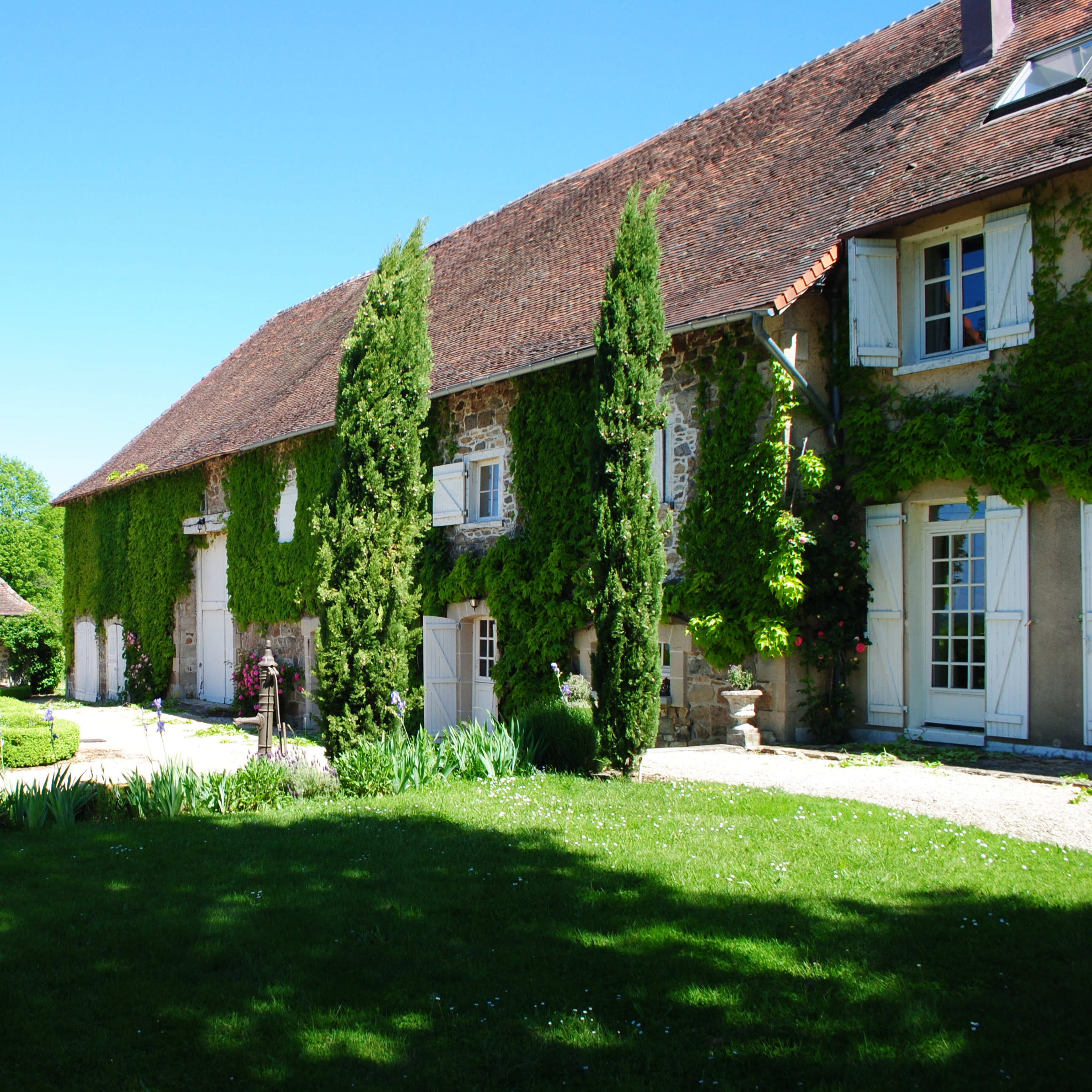 A charming stone farmhouse with a brown tiled roof, surrounded by lush greenery and a landscaped garden.