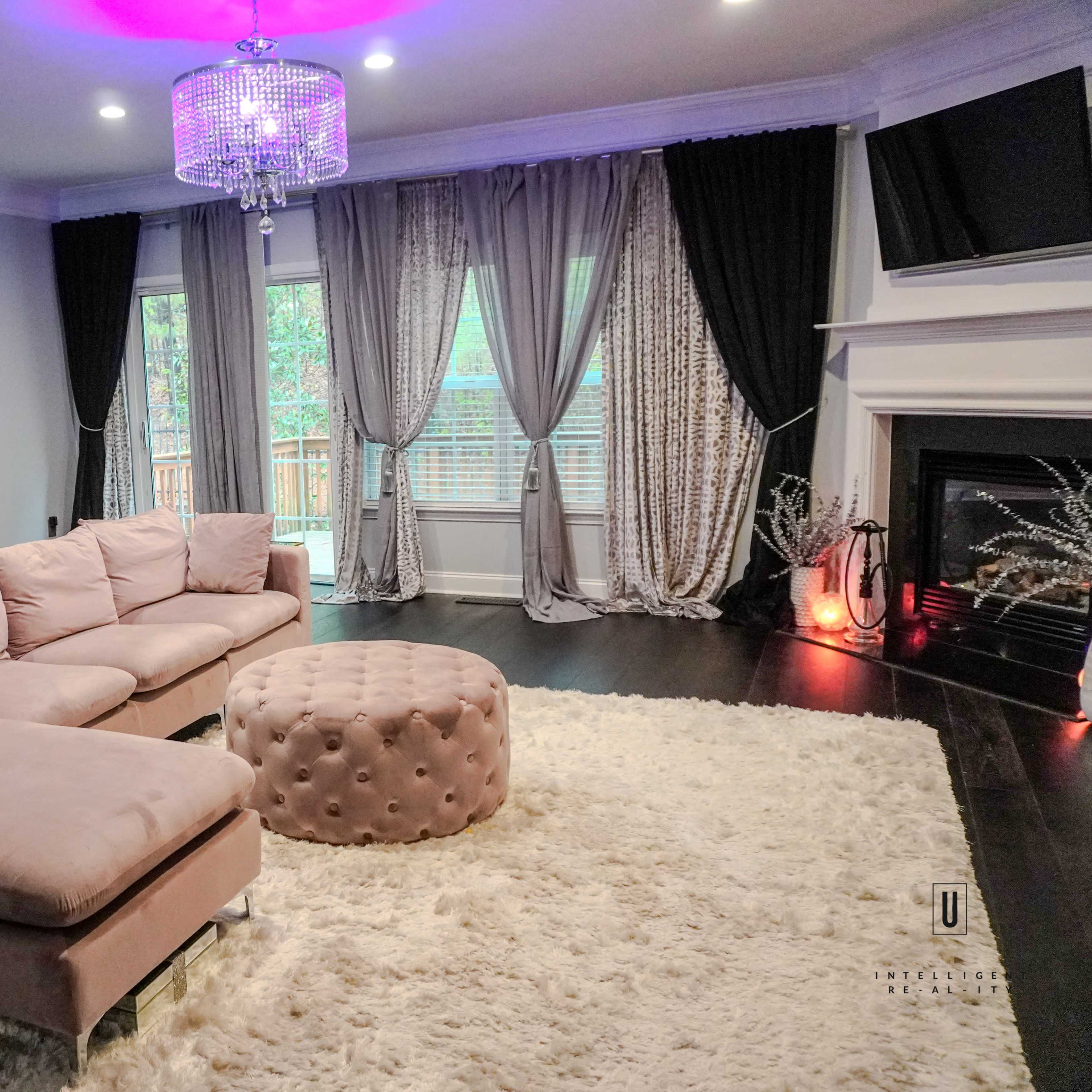 A living room featuring a pink sectional sofa, a tufted ottoman, a plush white rug, and large windows with curtains, all set against dark hardwood flooring.