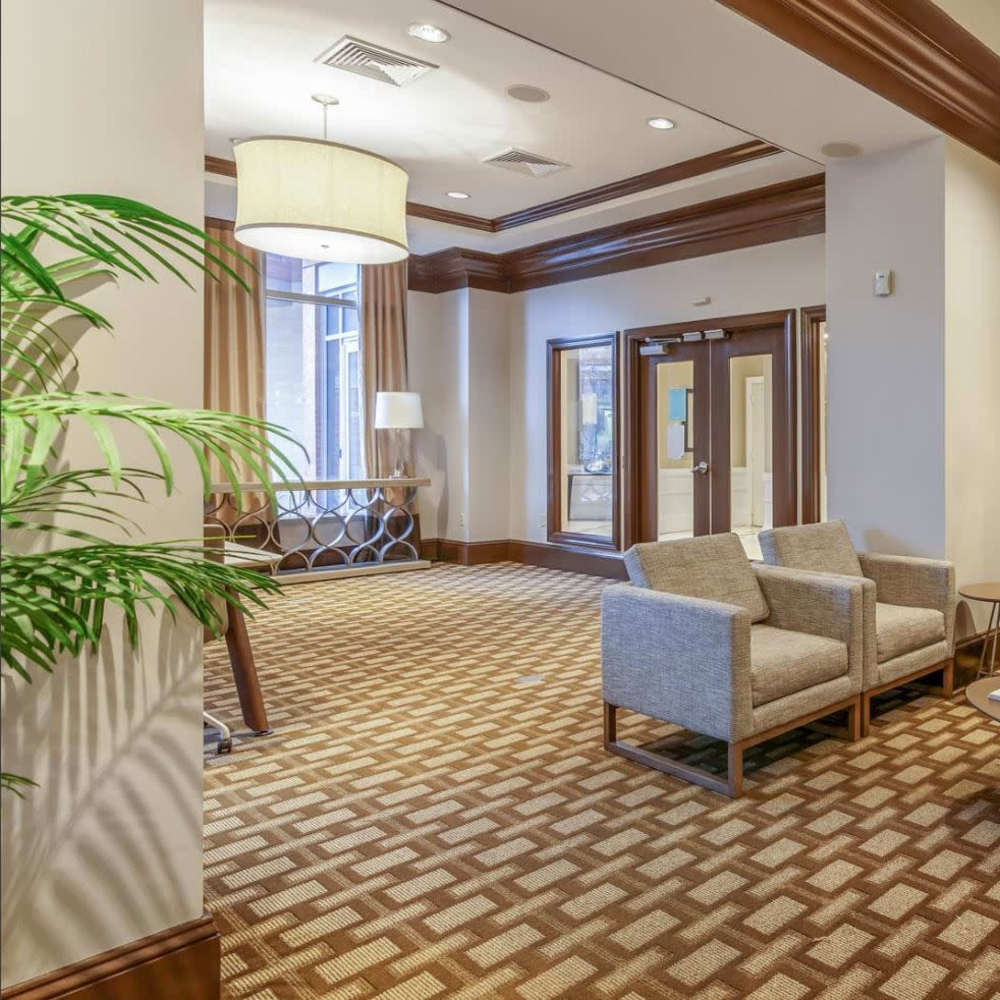 A modern lobby area featuring gray upholstered chairs, a wooden coffee table, and a decorative plant, with an entrance visible in the background.