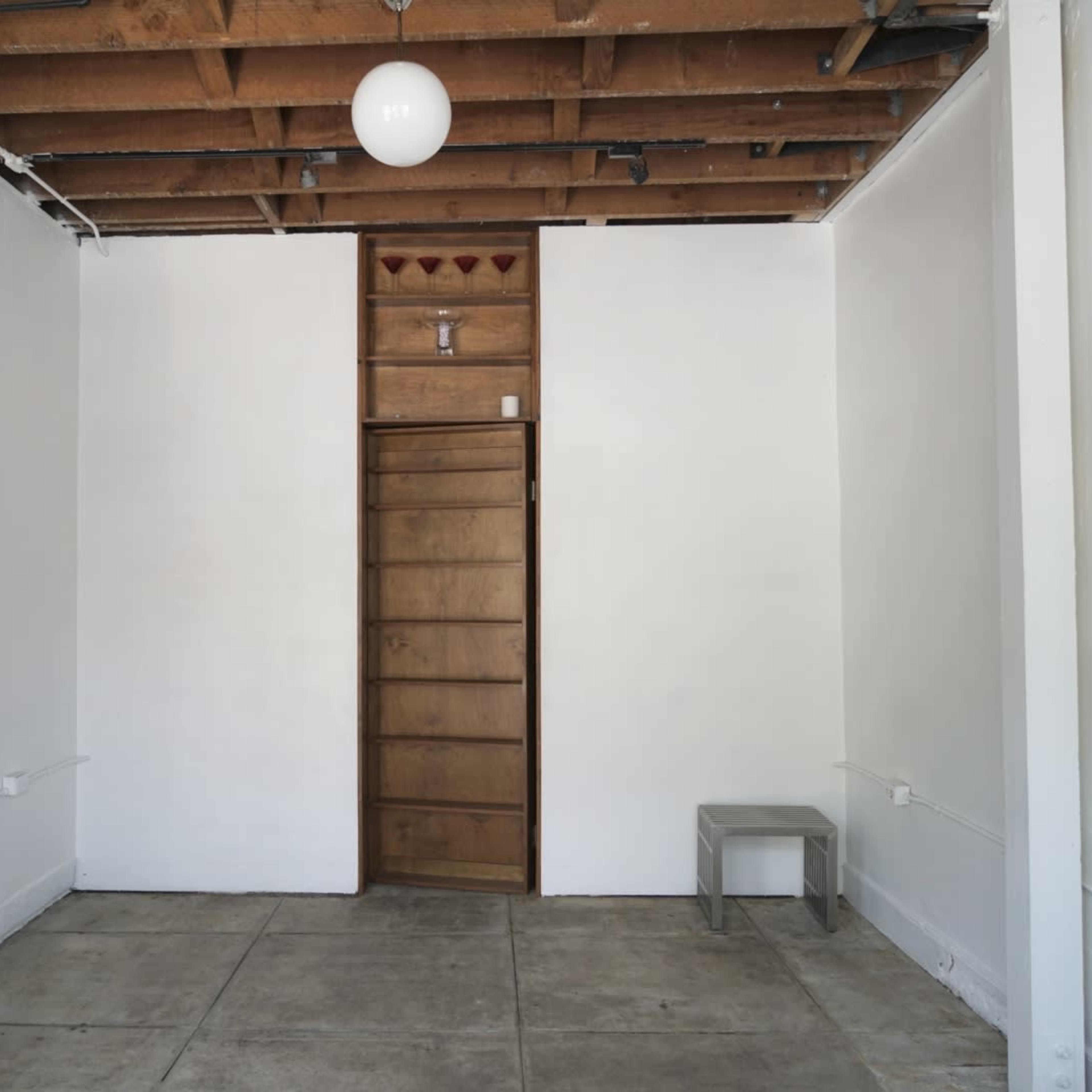A sparsely furnished room with exposed wooden beams, a single overhead light, a door at the far end, and a potted plant on the left wall.
