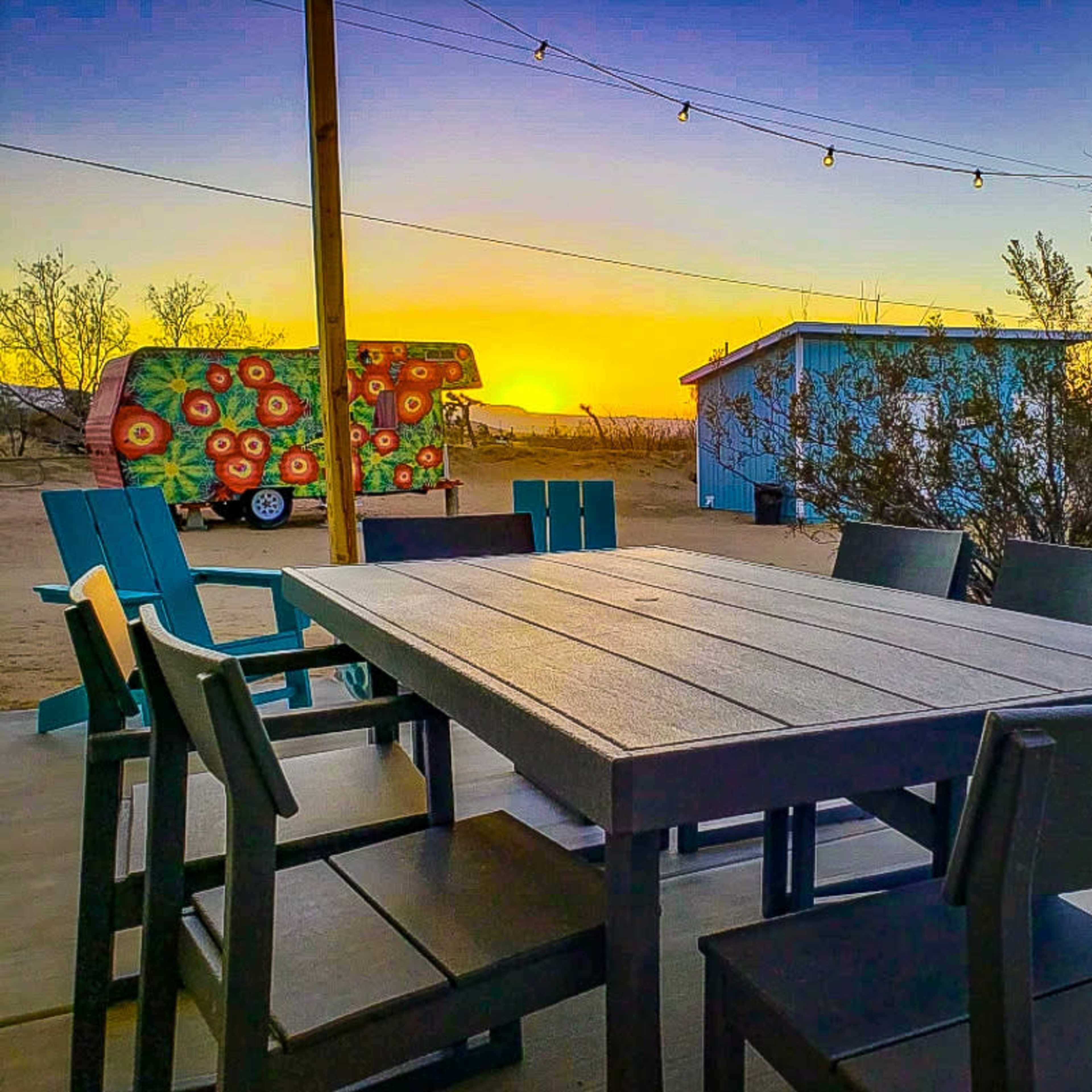 A wooden dining table with chairs is set in the foreground, facing a colorful food truck and a blue building against a sunset in the background.