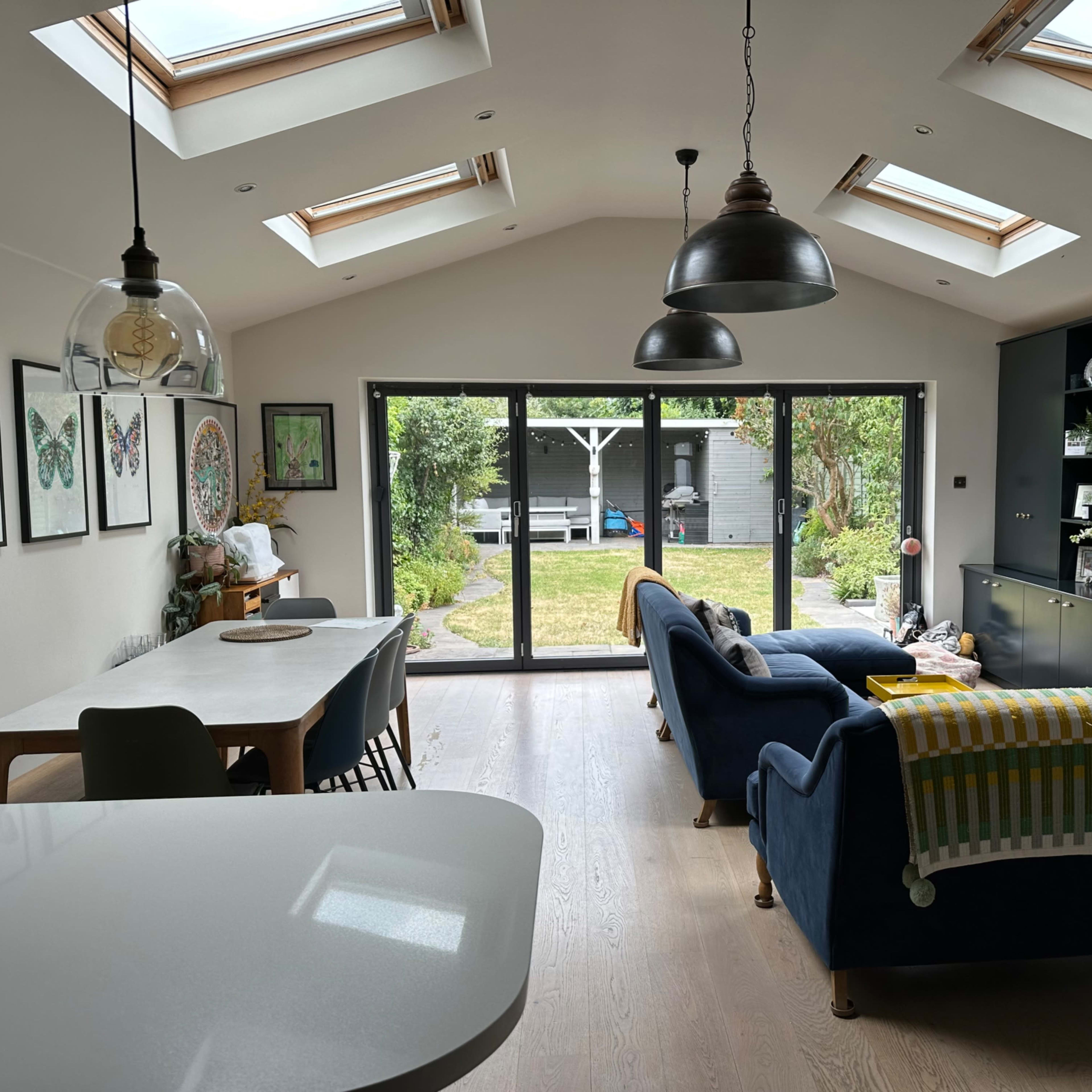 A modern living space with a light-filled interior featuring a dining area, a seating area with blue sofas, and large glass doors opening to a garden.