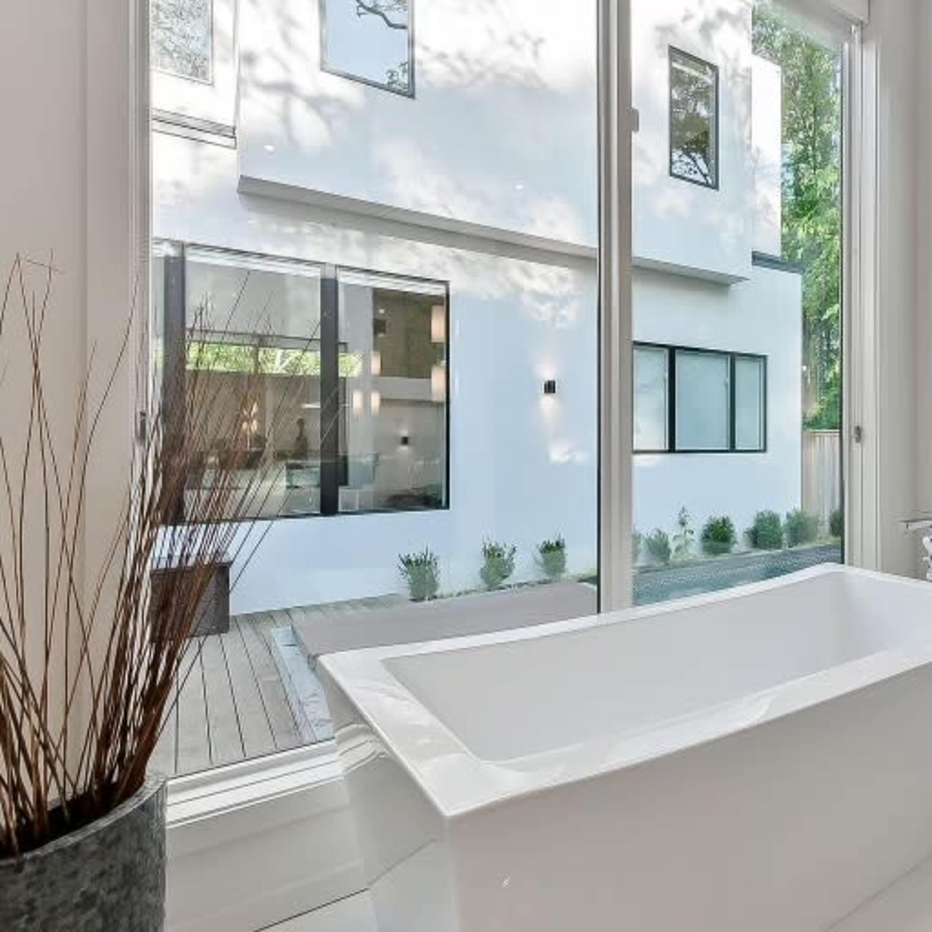 A freestanding bathtub is positioned next to large windows overlooking a green courtyard with patio seating.