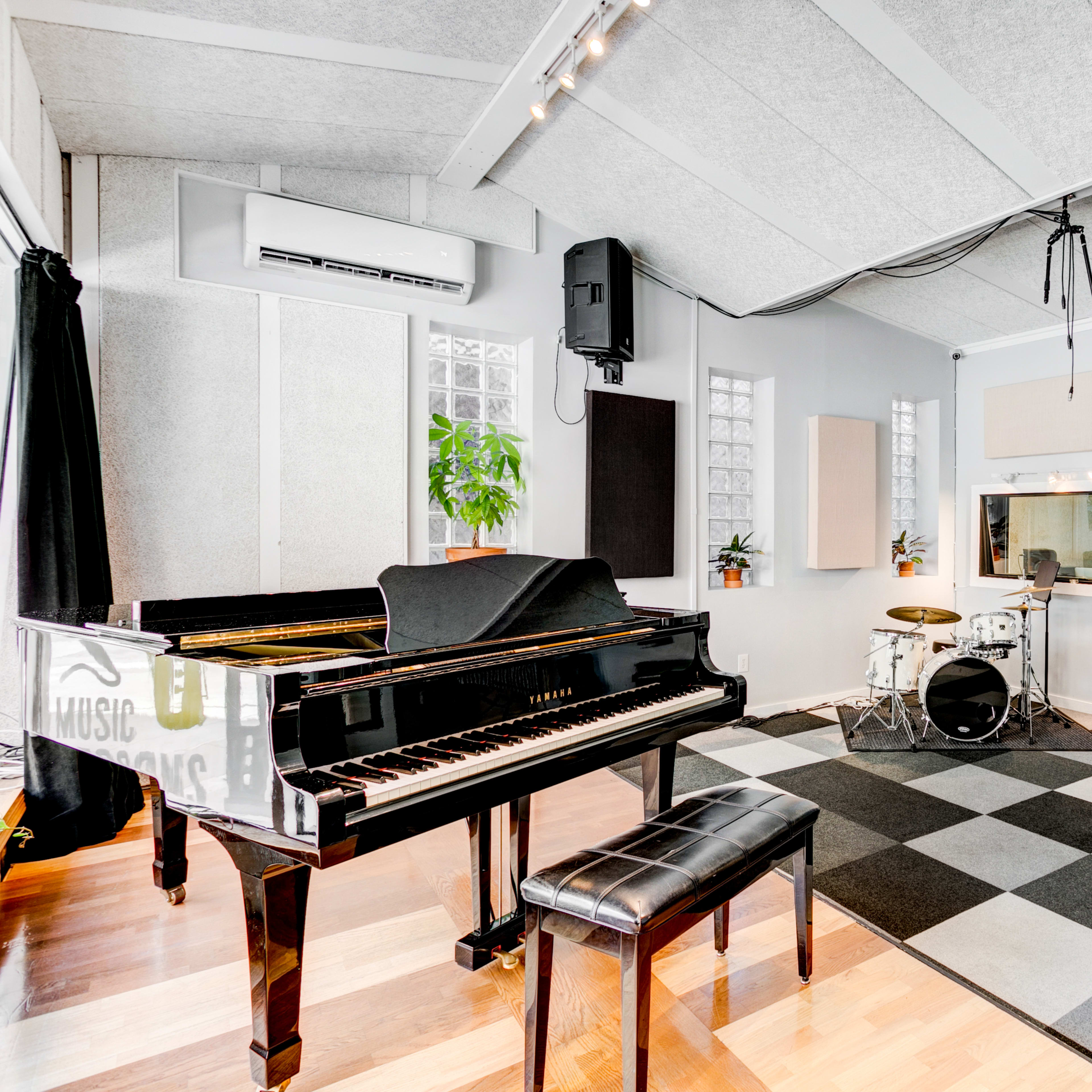 A music studio featuring a grand piano, a drum set, and various guitars displayed on the walls, with a checkerboard floor and large windows.