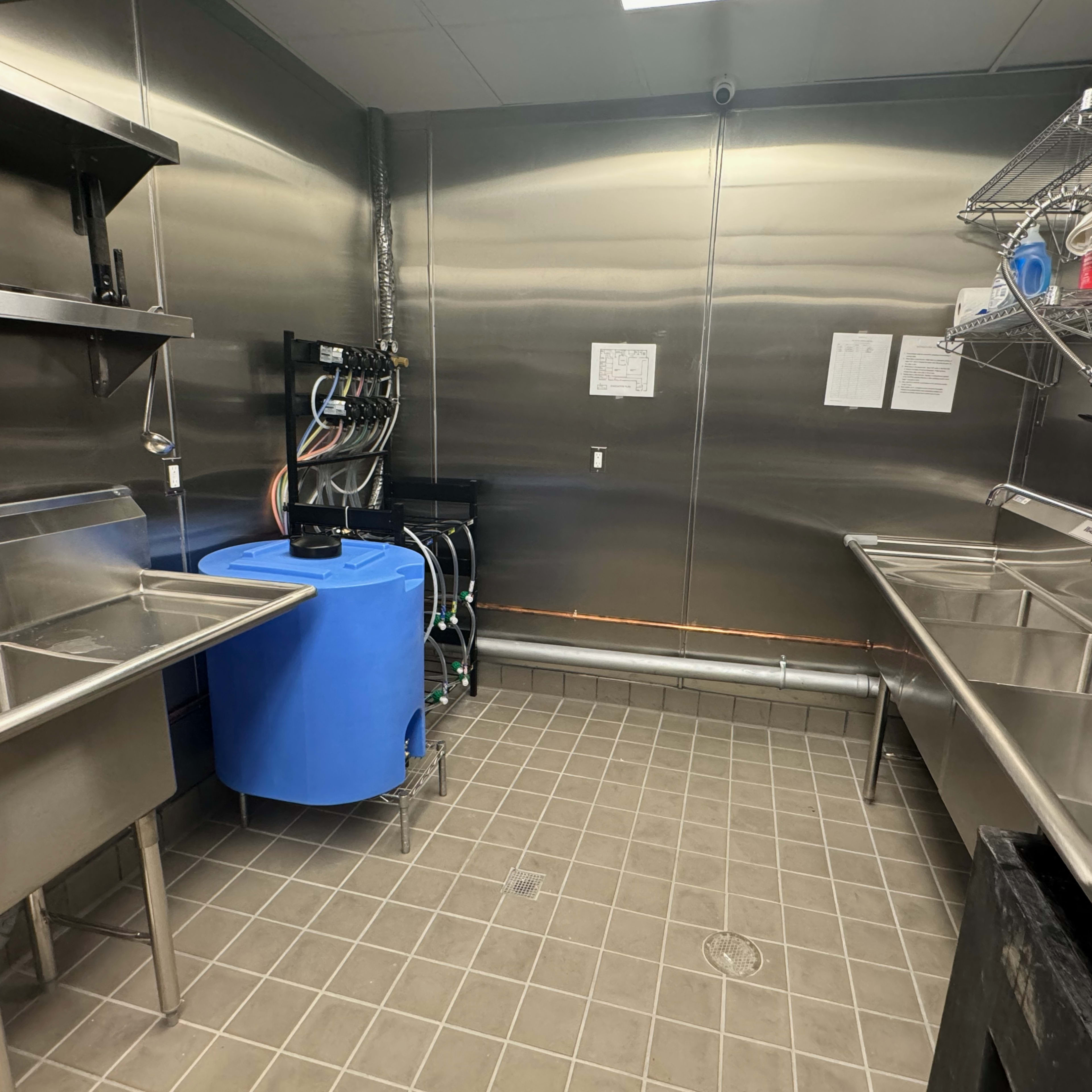 A stainless steel kitchen area equipped with several sinks, a large blue water tank, and metal shelving units.