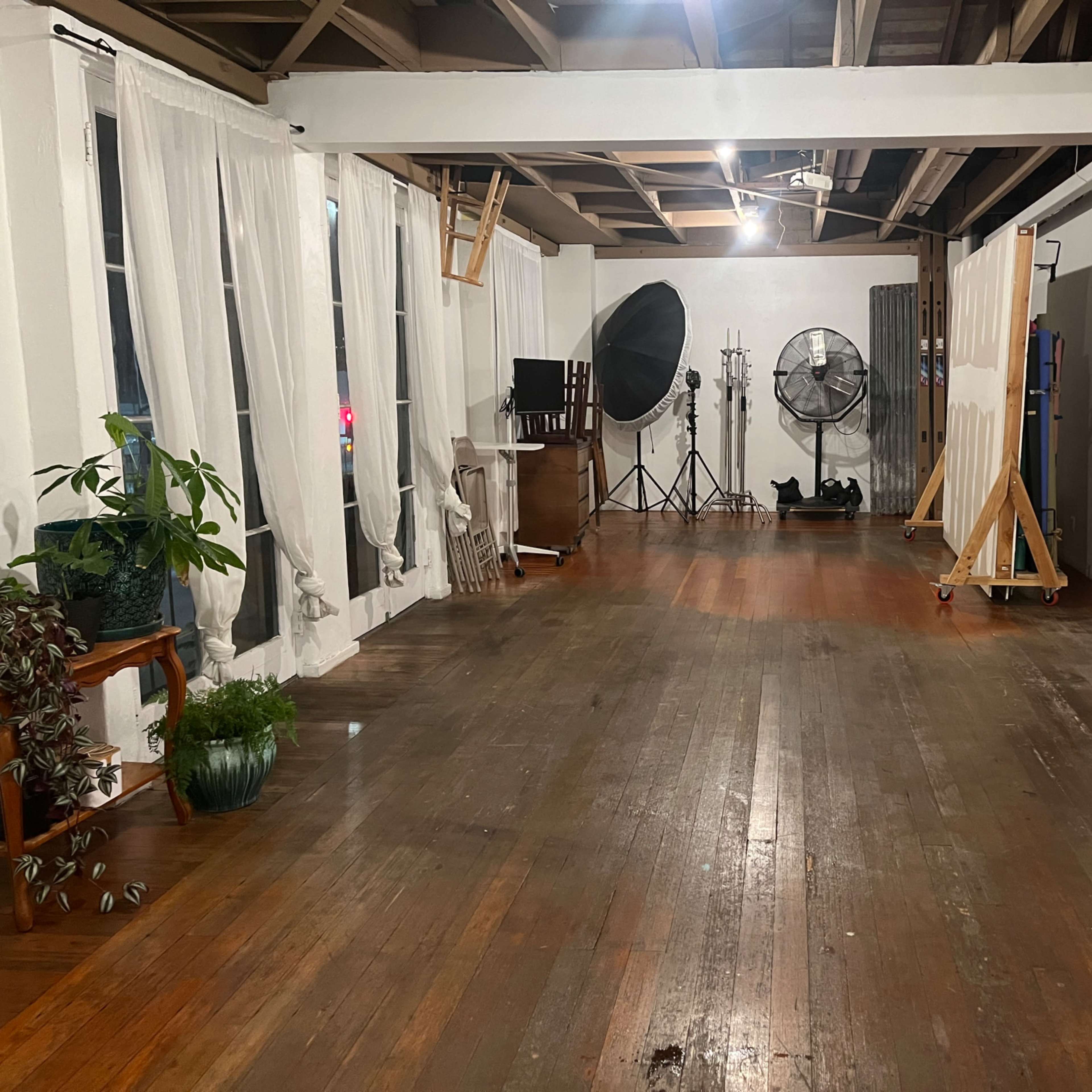 A spacious room with wooden floors, large windows covered by white curtains, and various studio equipment and a dog in one corner.