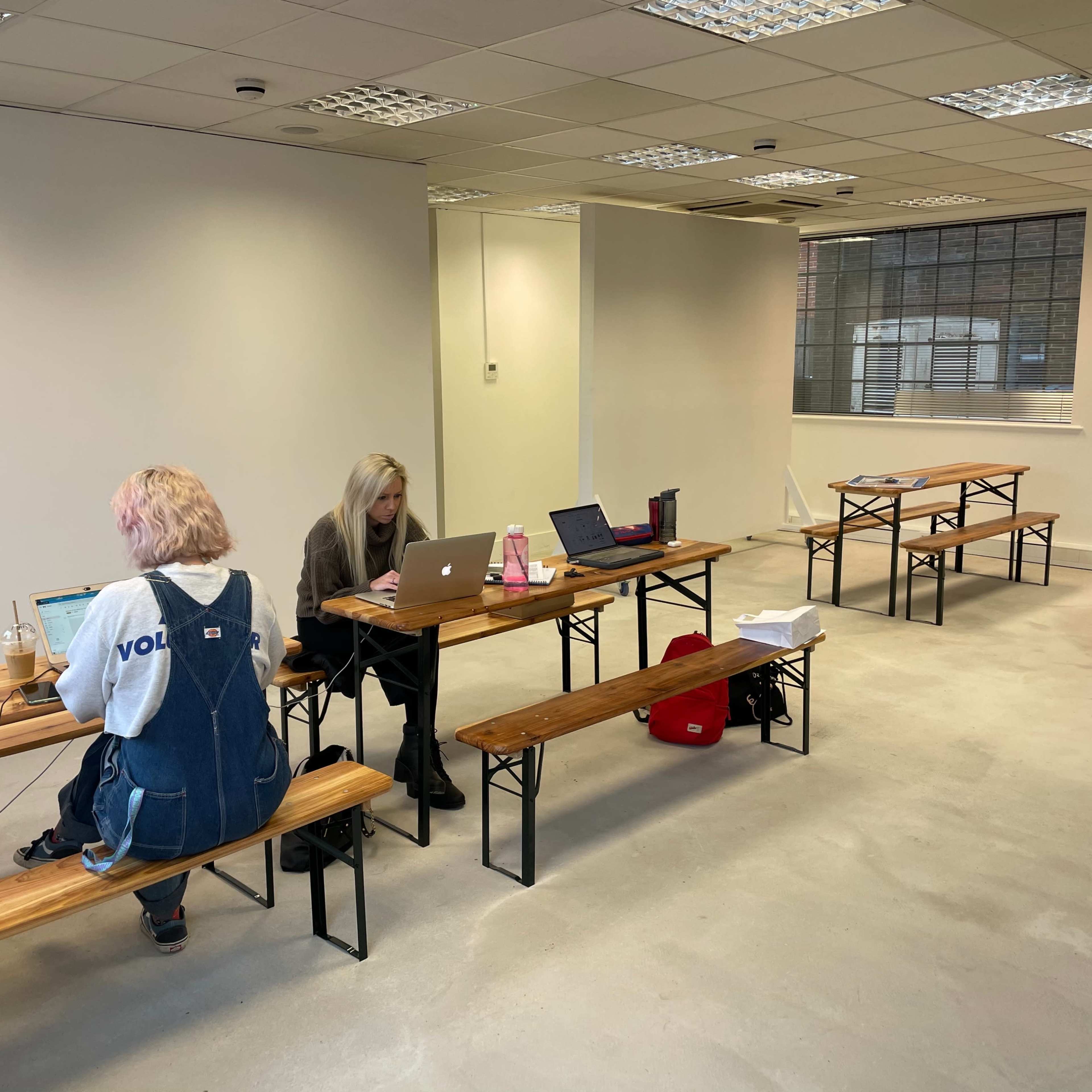 Two people work on laptops at wooden tables in a sparsely furnished room with large windows.