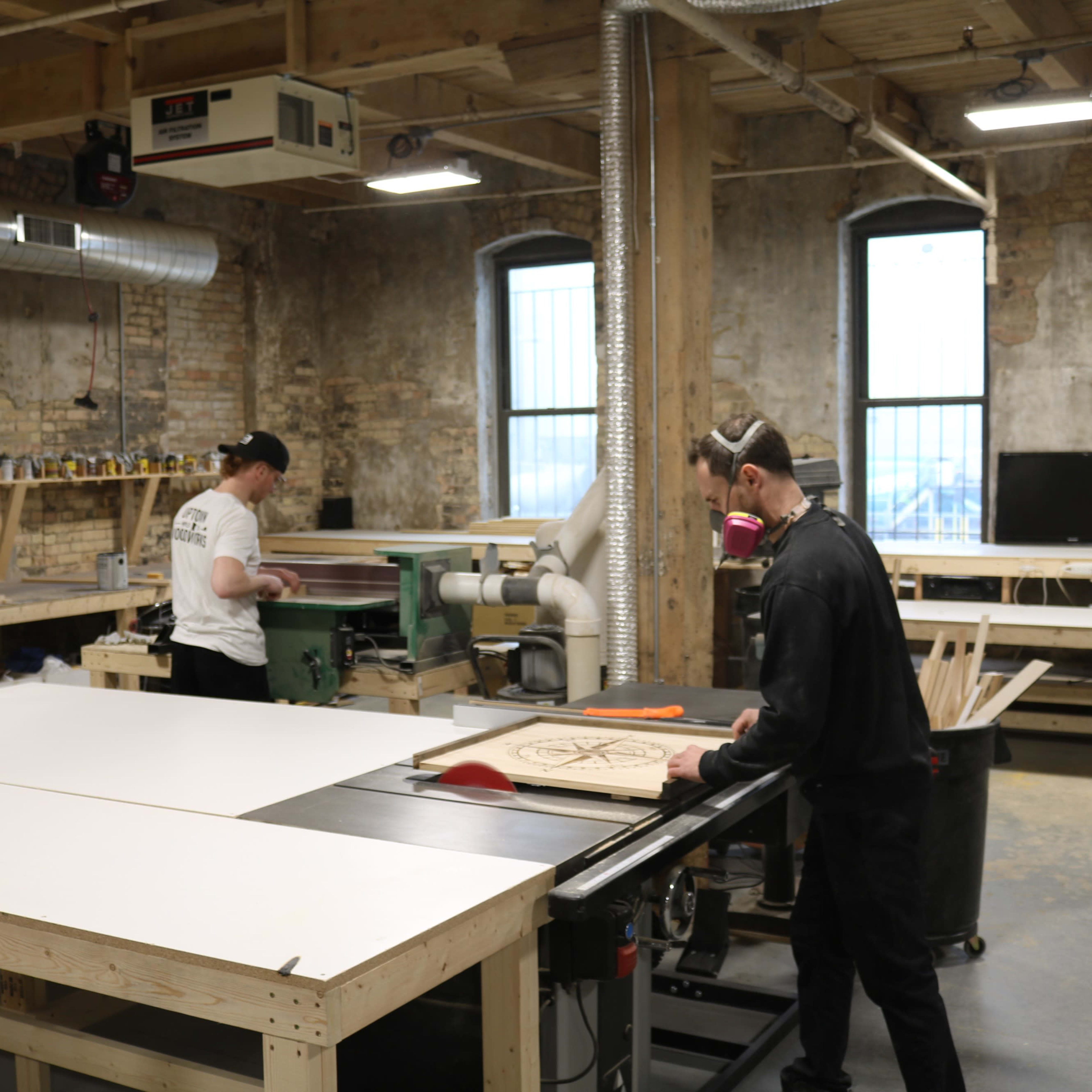 Two individuals are working in a woodshop, one operating a table saw and the other using a planer.