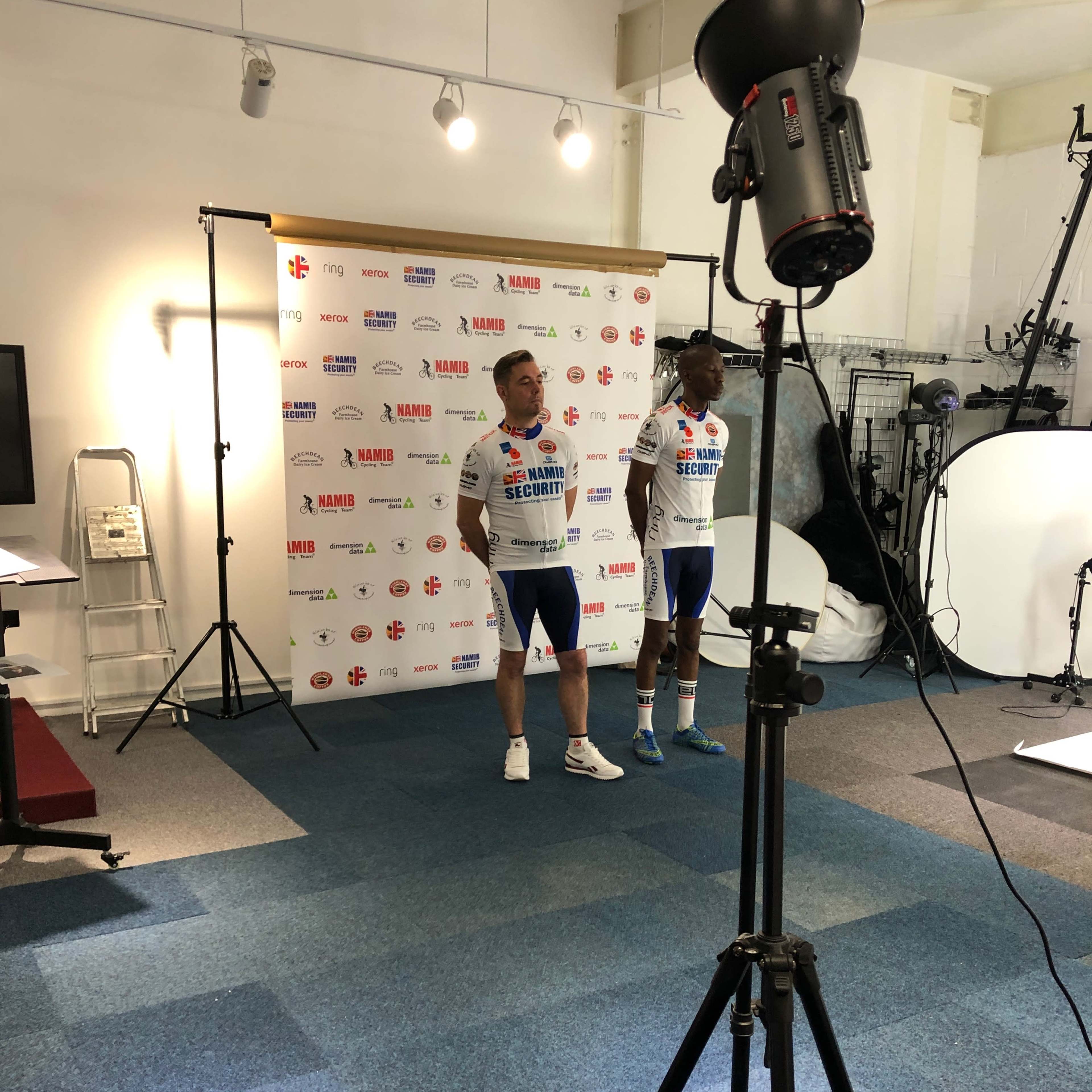 Two cyclists in team jerseys stand in front of a sponsor logo backdrop during a photoshoot in a studio setting.