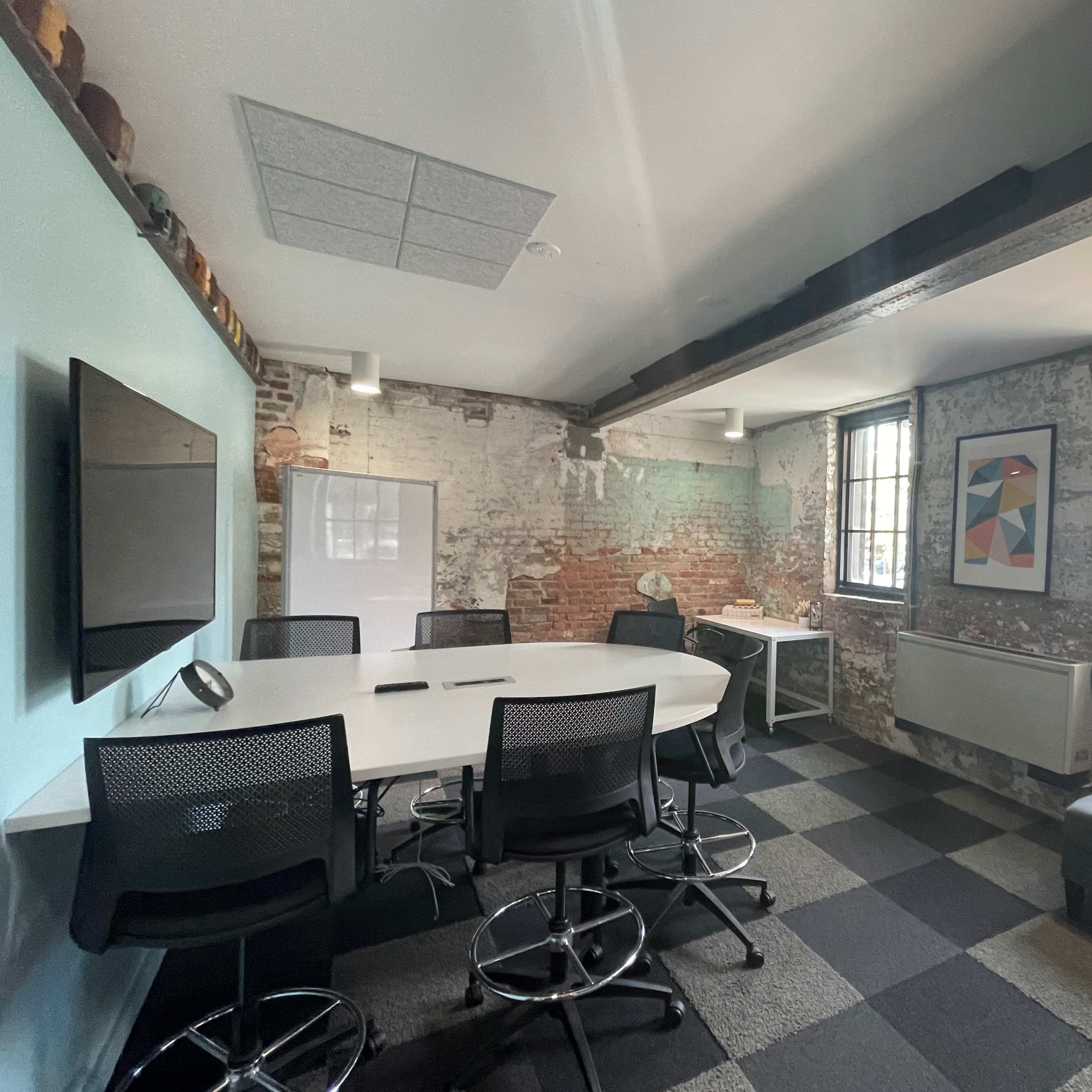 A modern conference room with a round table surrounded by black swivel chairs, exposed brick walls, a large screen, and two windows allowing natural light.