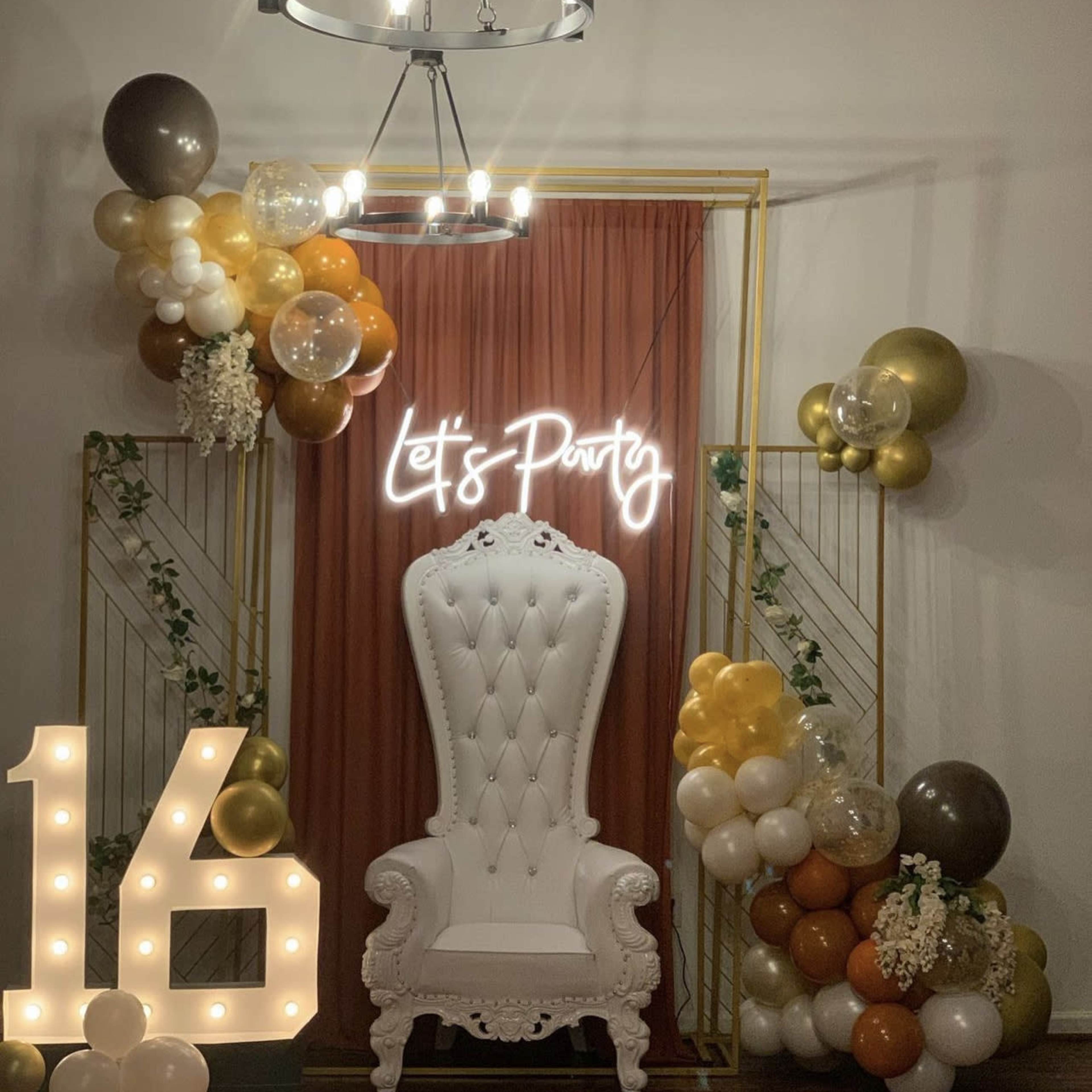 A decorated party space with a white throne chair, a neon sign reading "Let's Party," gold and brown balloons, and illuminated numbers reading "16."