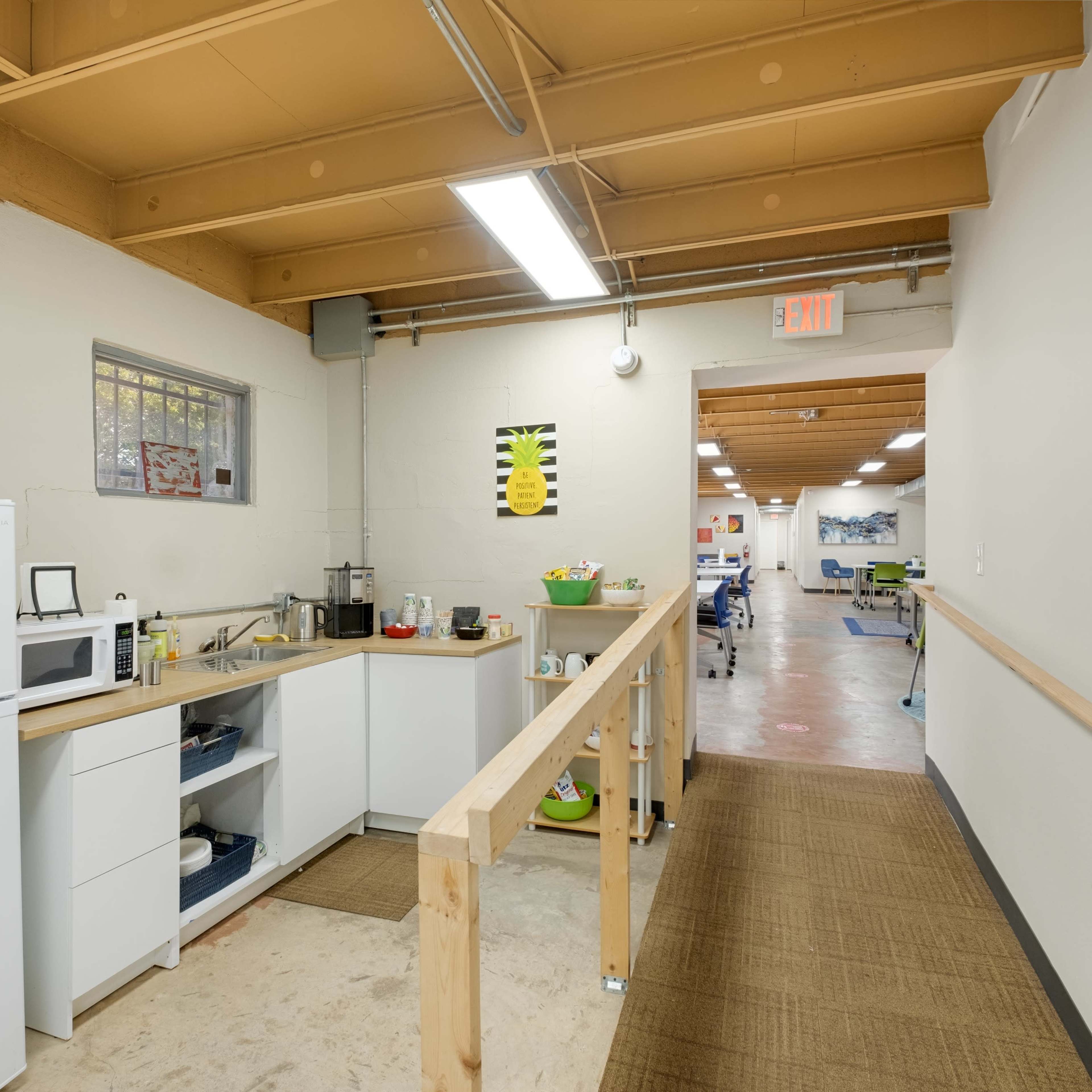 A narrow hallway leading to a kitchen area with a refrigerator, microwave, and counter space, along with a ramp and an open area beyond.