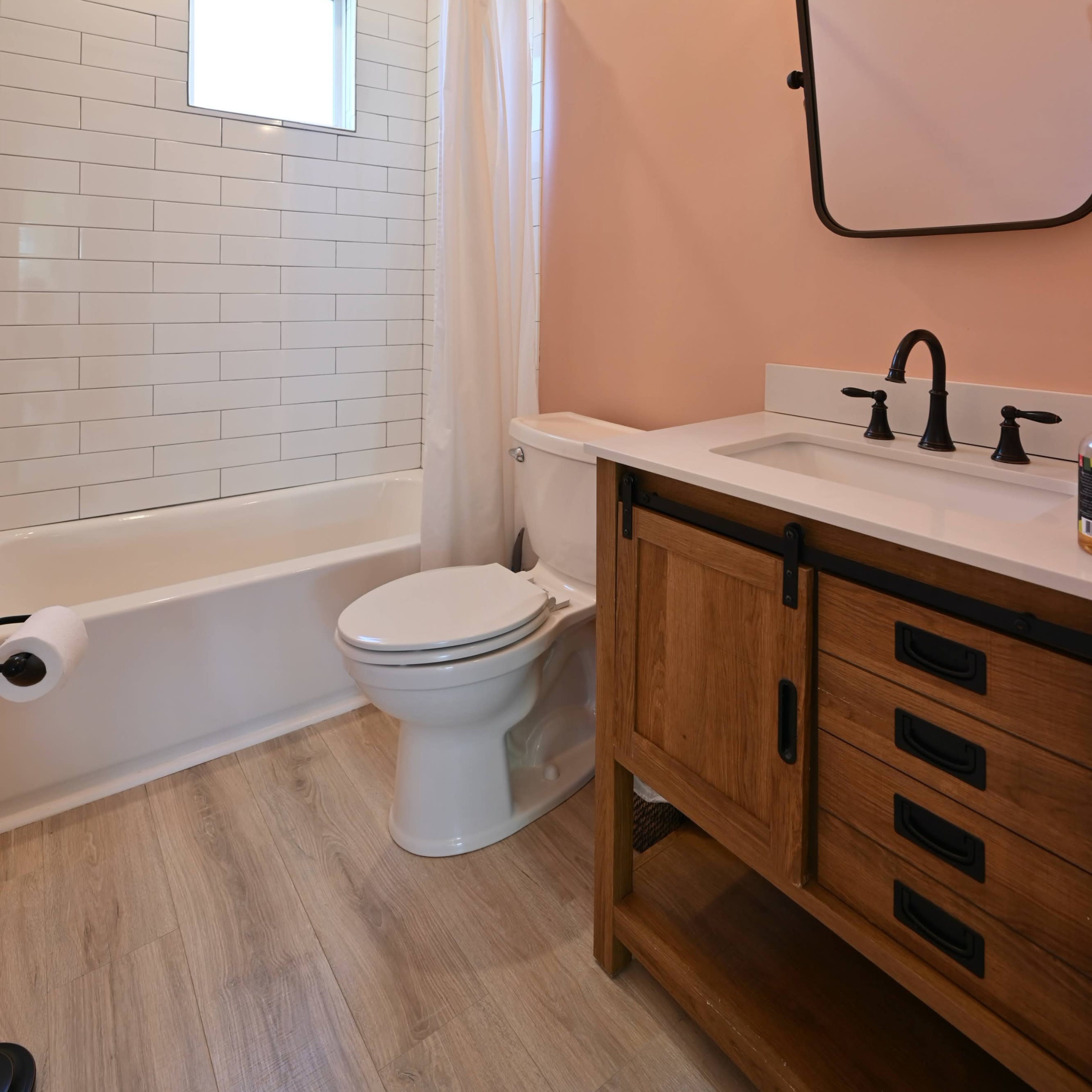 A bathroom featuring a bathtub with a shower, a toilet, a wooden vanity, a mirror, and light fixtures against a pink wall.