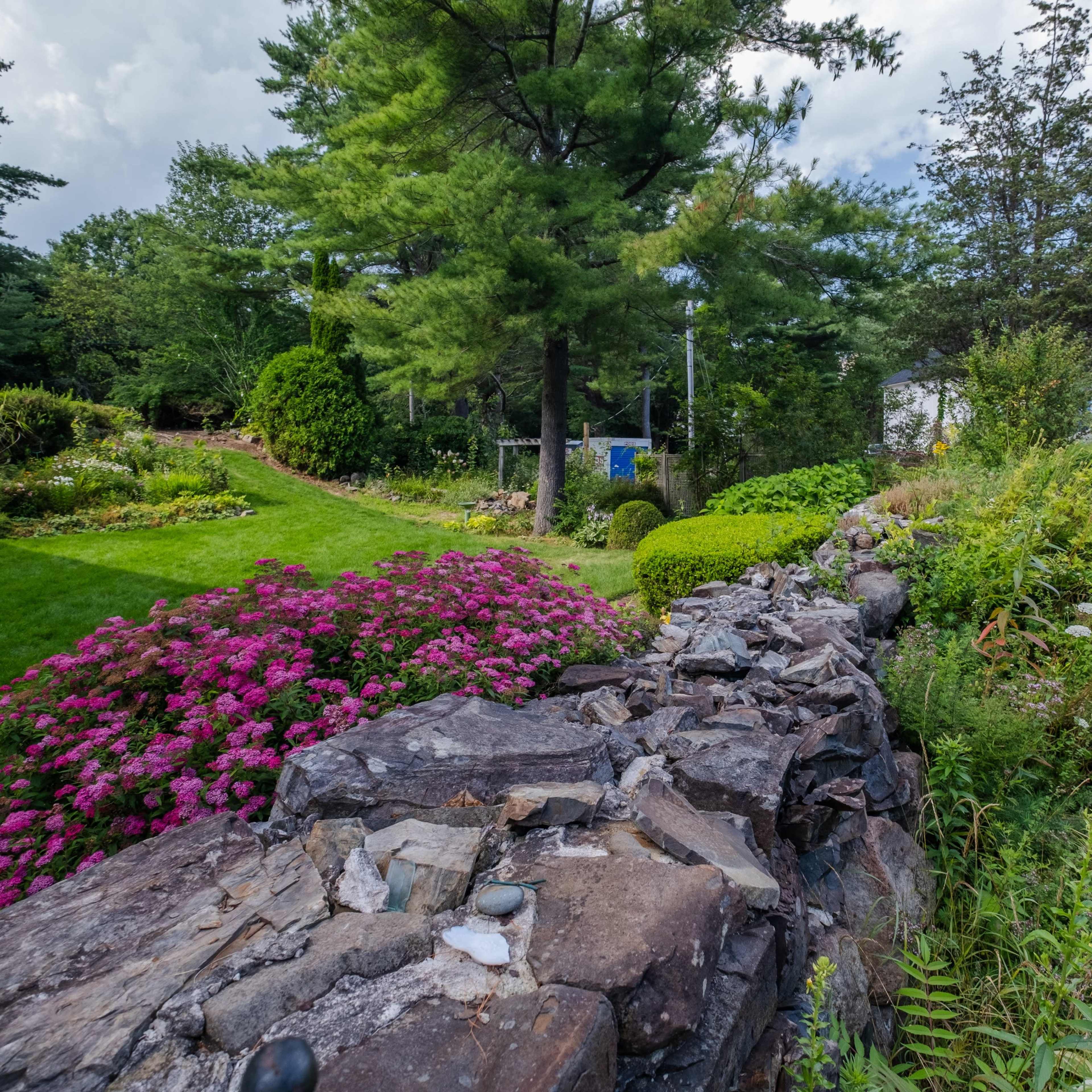 A landscaped garden featuring a stone wall, colorful flower beds, and tall trees under a partly cloudy sky.