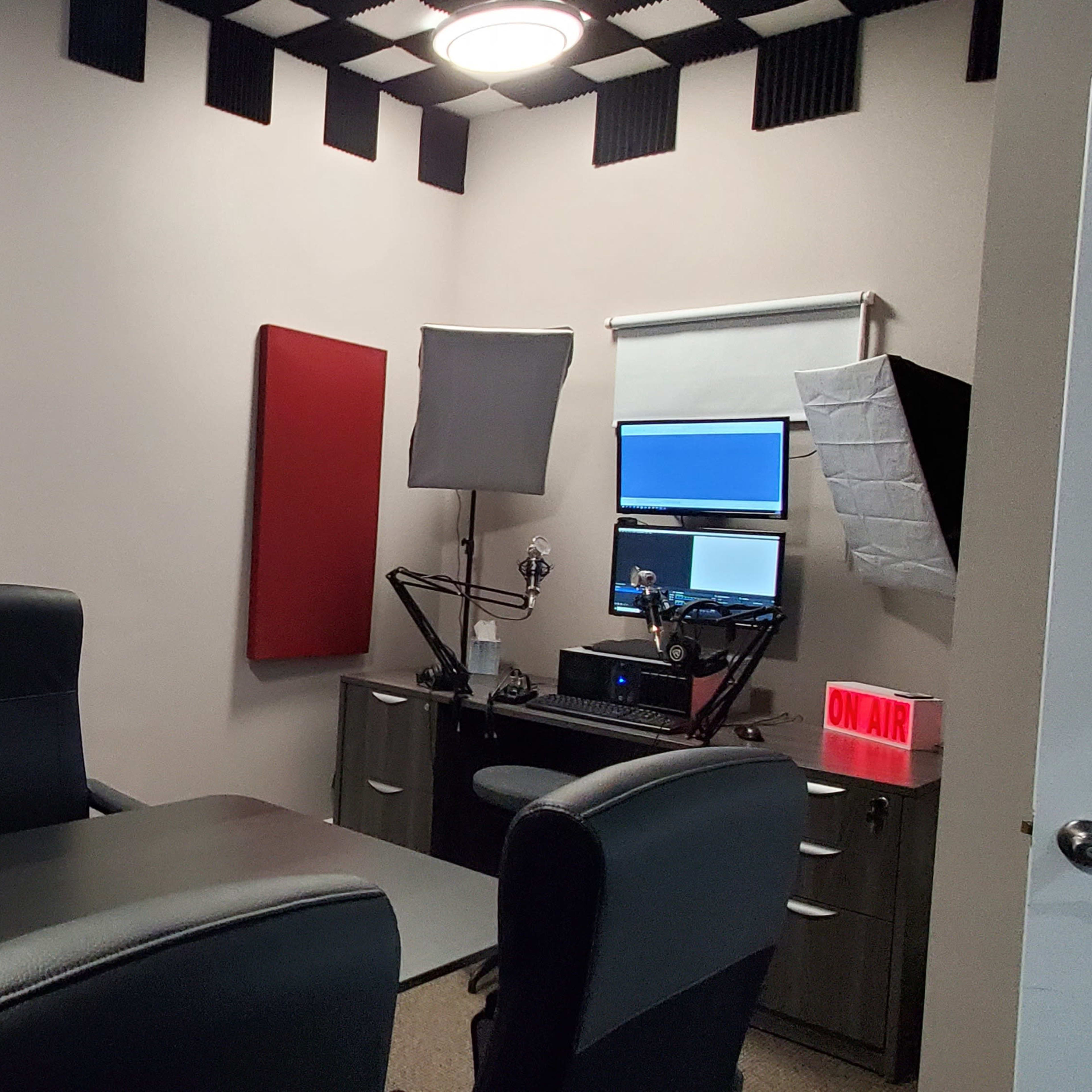 A recording studio with a long table and three black chairs, equipped with a computer, microphone, and soundproofing panels.