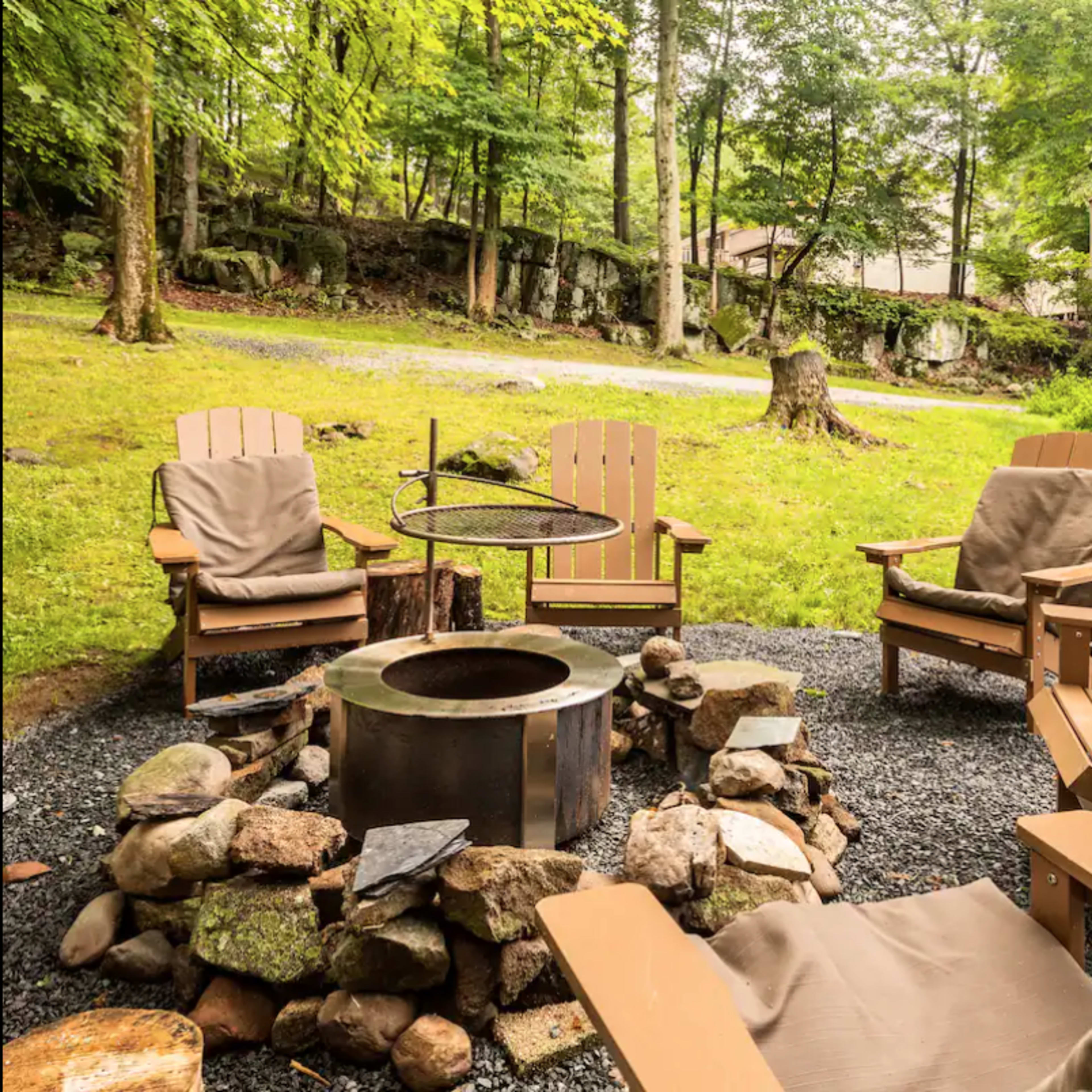 A circular fire pit surrounded by wooden chairs is set in a grassy area near a house and trees.