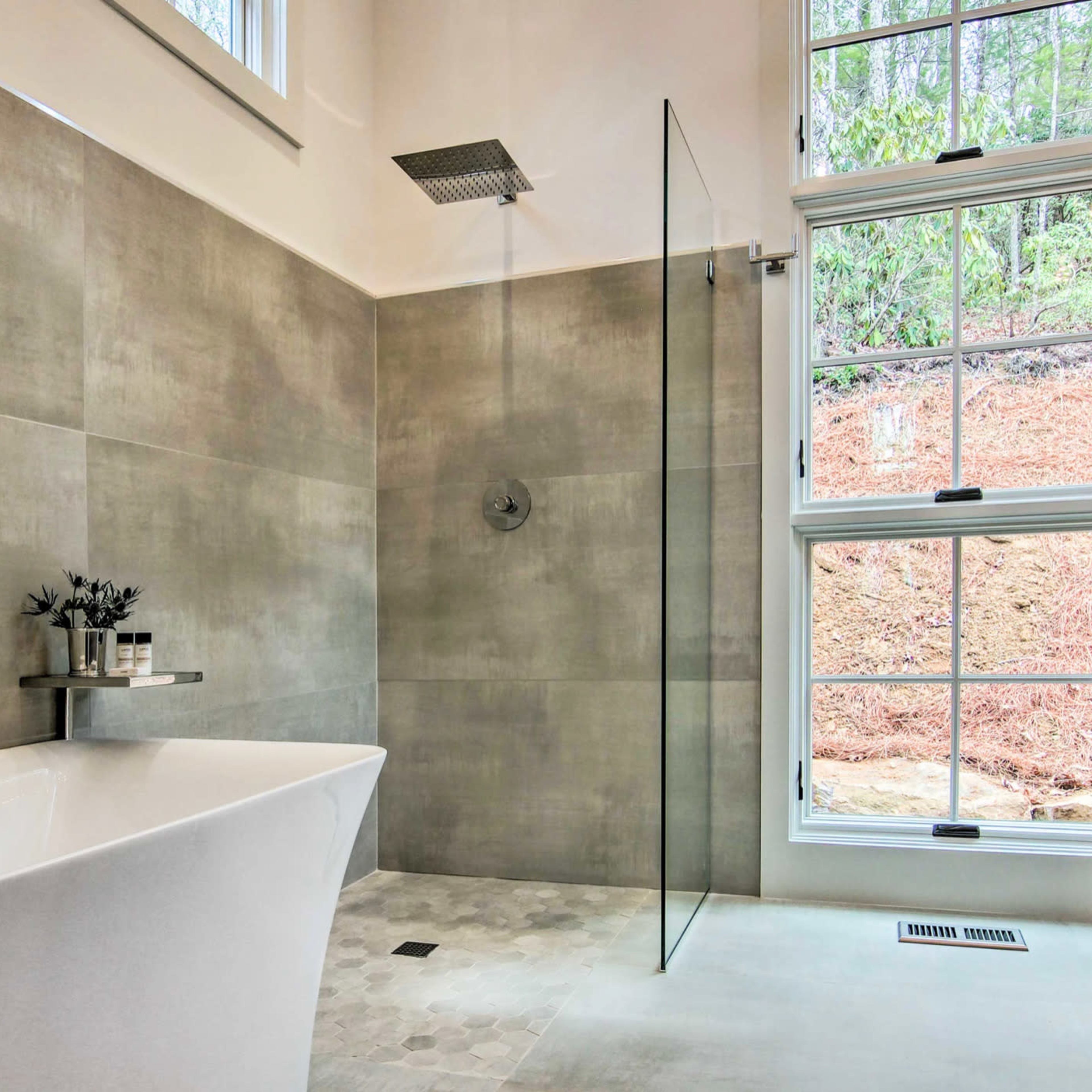 A modern bathroom featuring a freestanding bathtub, a glass shower enclosure, and large windows with a view of greenery outside.