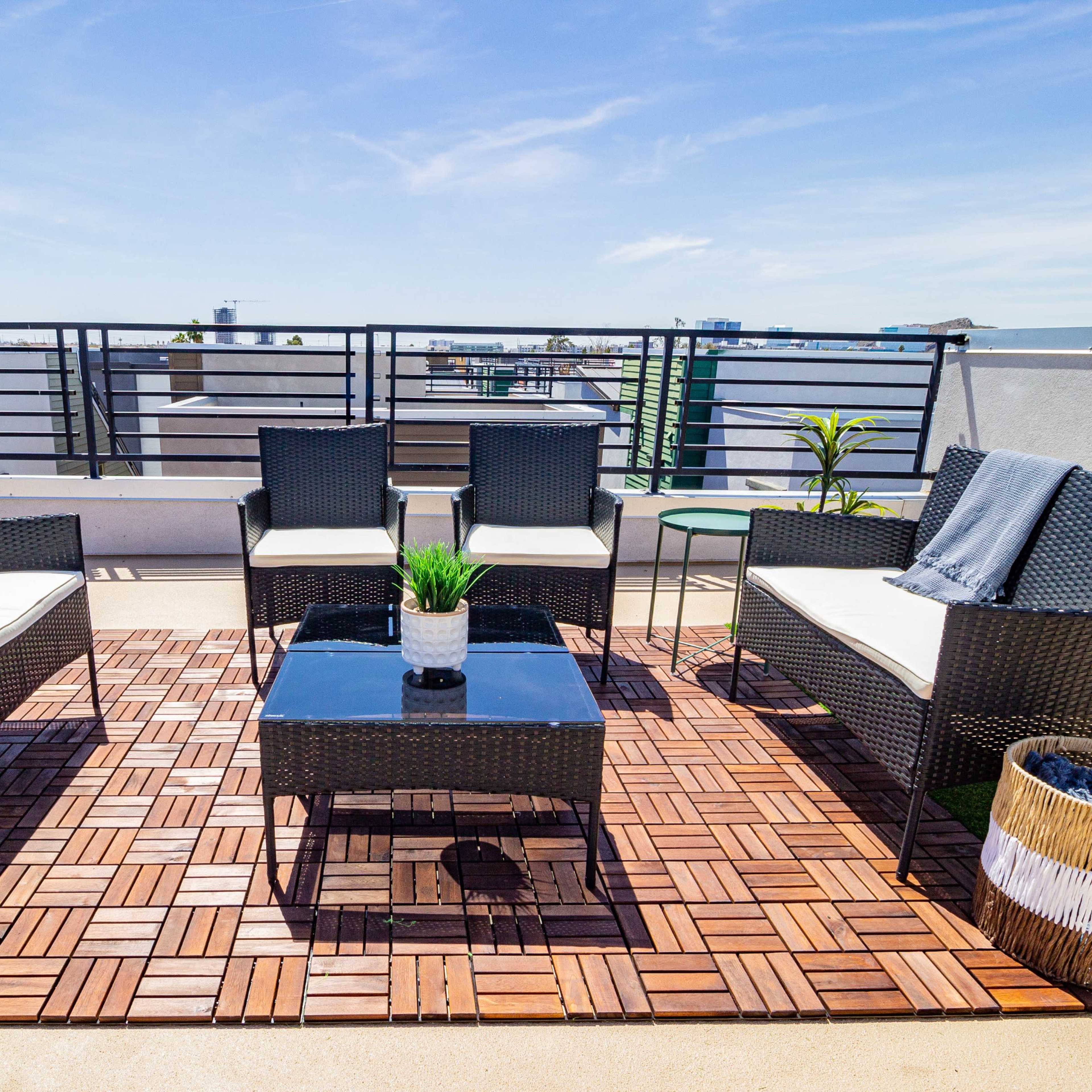 A rooftop patio with four wicker chairs arranged around a black table on a wooden deck, with a potted plant and a decorative basket nearby.