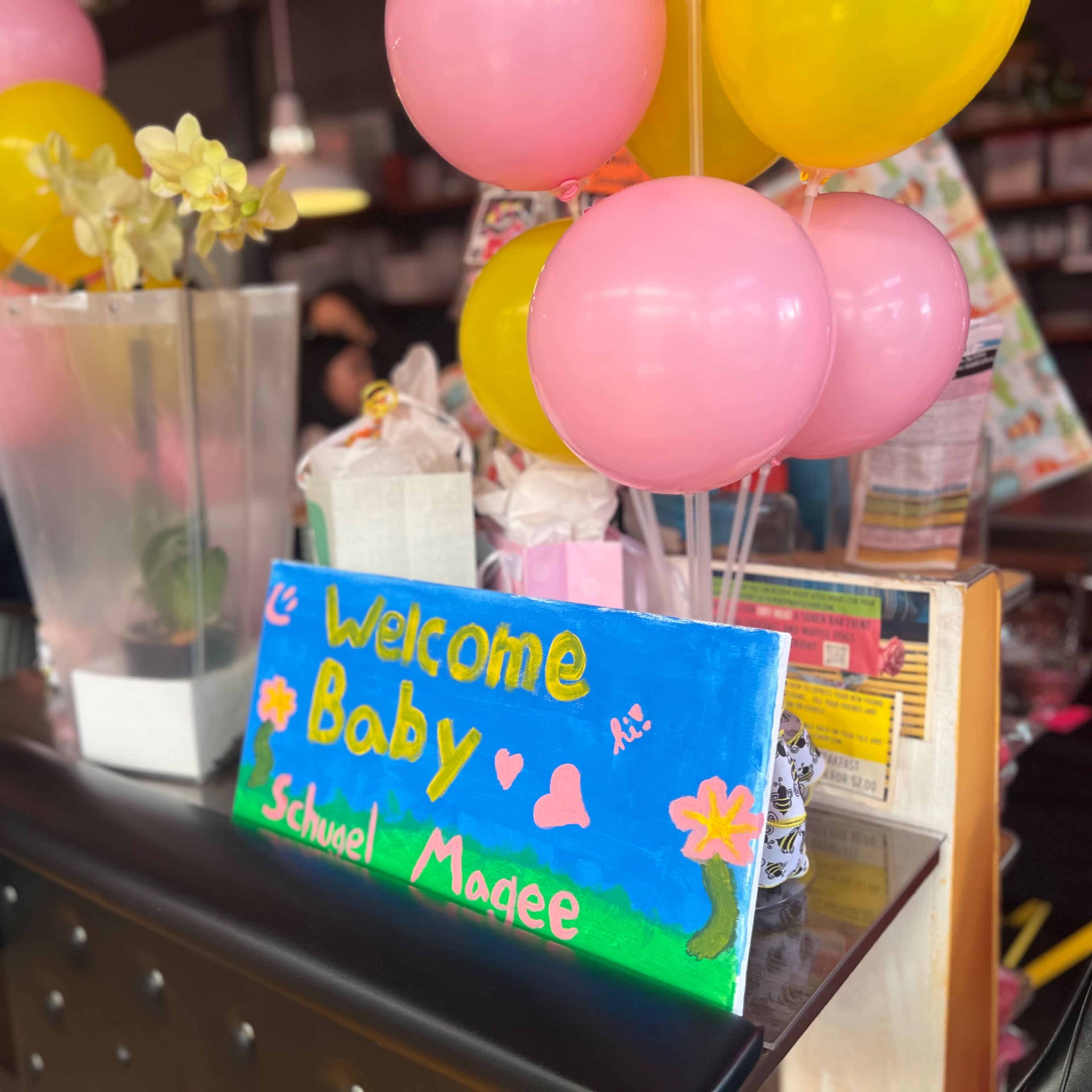 The scene features a colorful welcome sign for a baby, surrounded by balloons and decorations at a festive gathering.