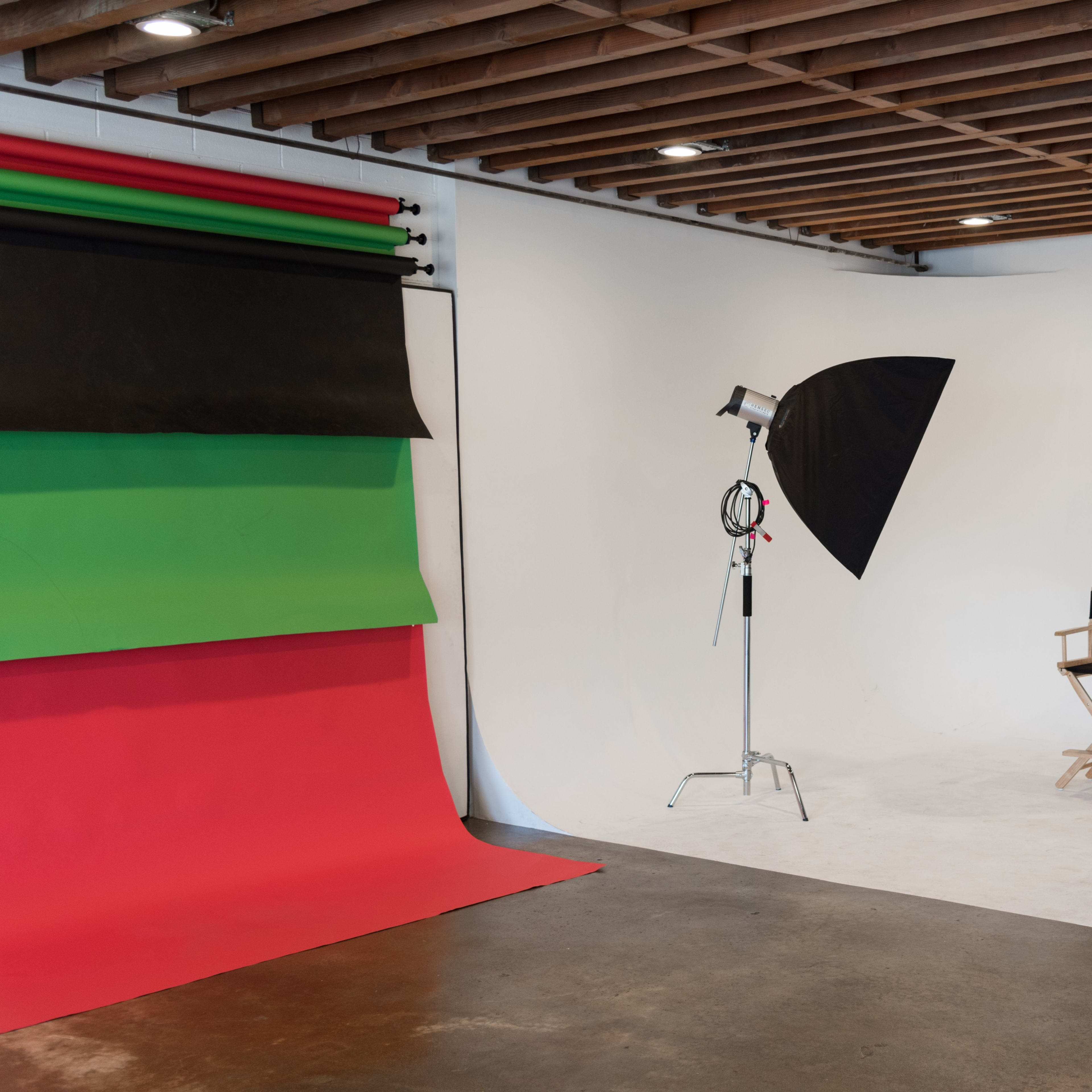 A photography studio with a green and red backdrop, two lighting setups, and a director's chair against a white wall.