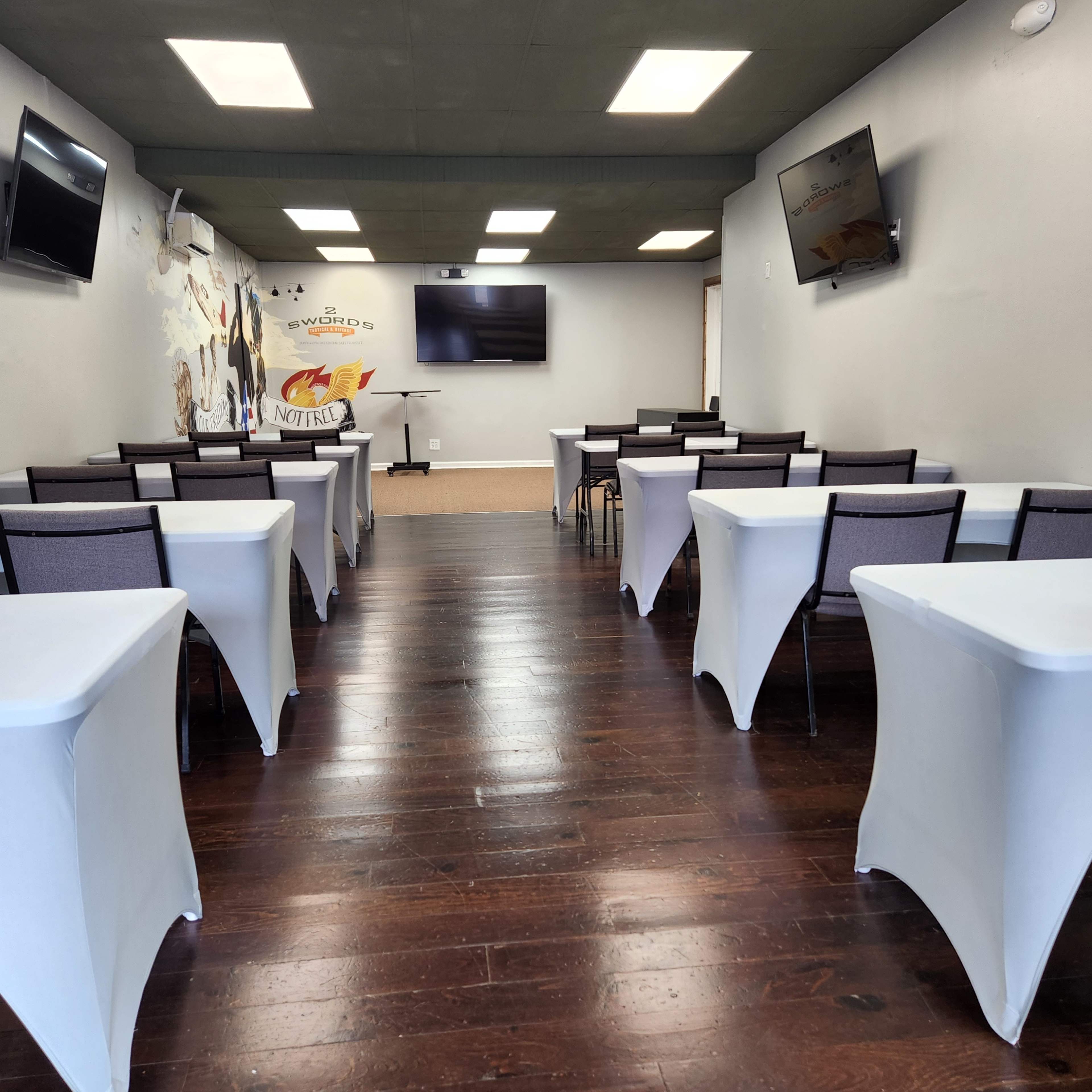 A classroom or meeting space with rows of tables covered in white tablecloths, equipped with wall-mounted televisions and a mural on one wall.