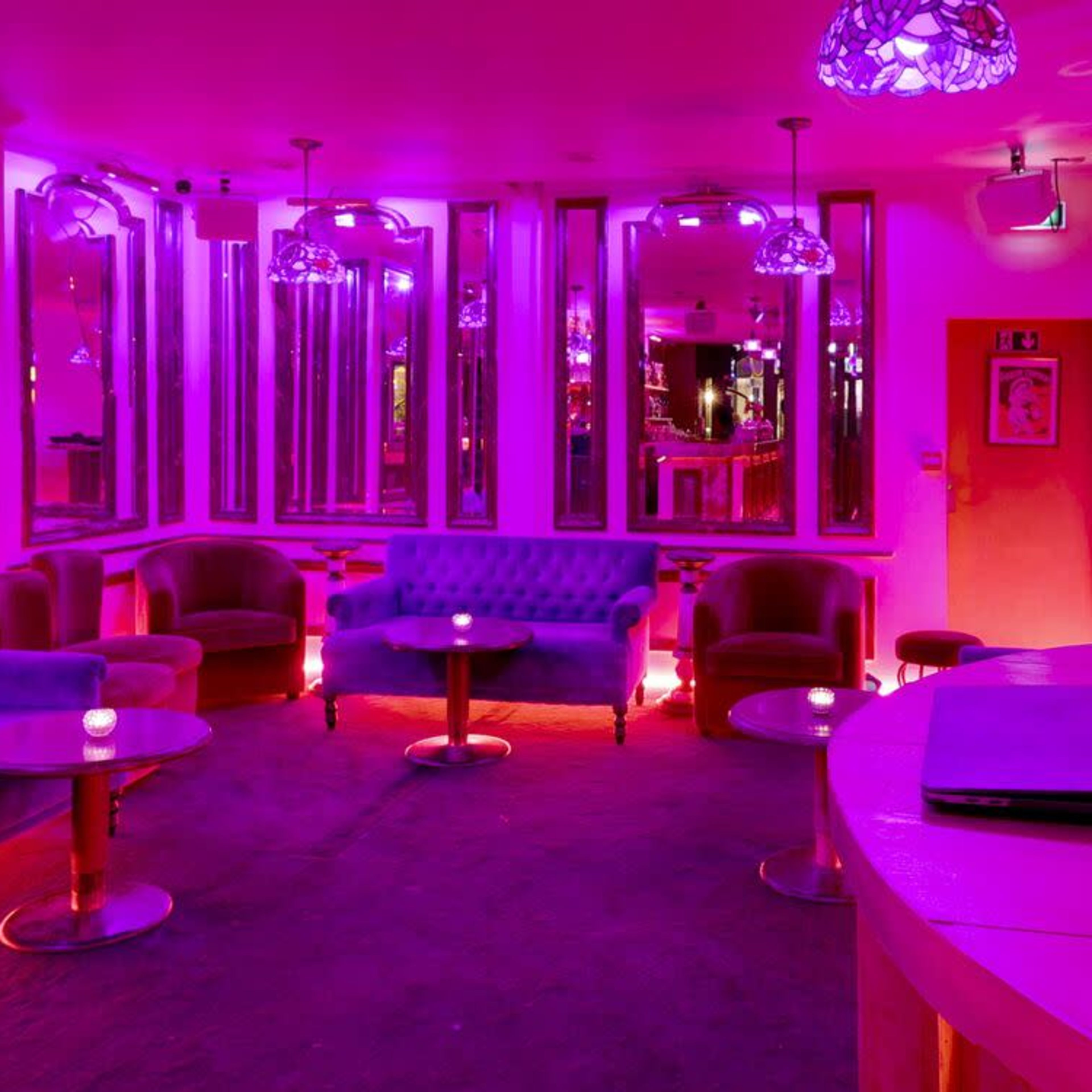 A lounge area with purple lighting features plush seating, mirrored walls, and a DJ setup.