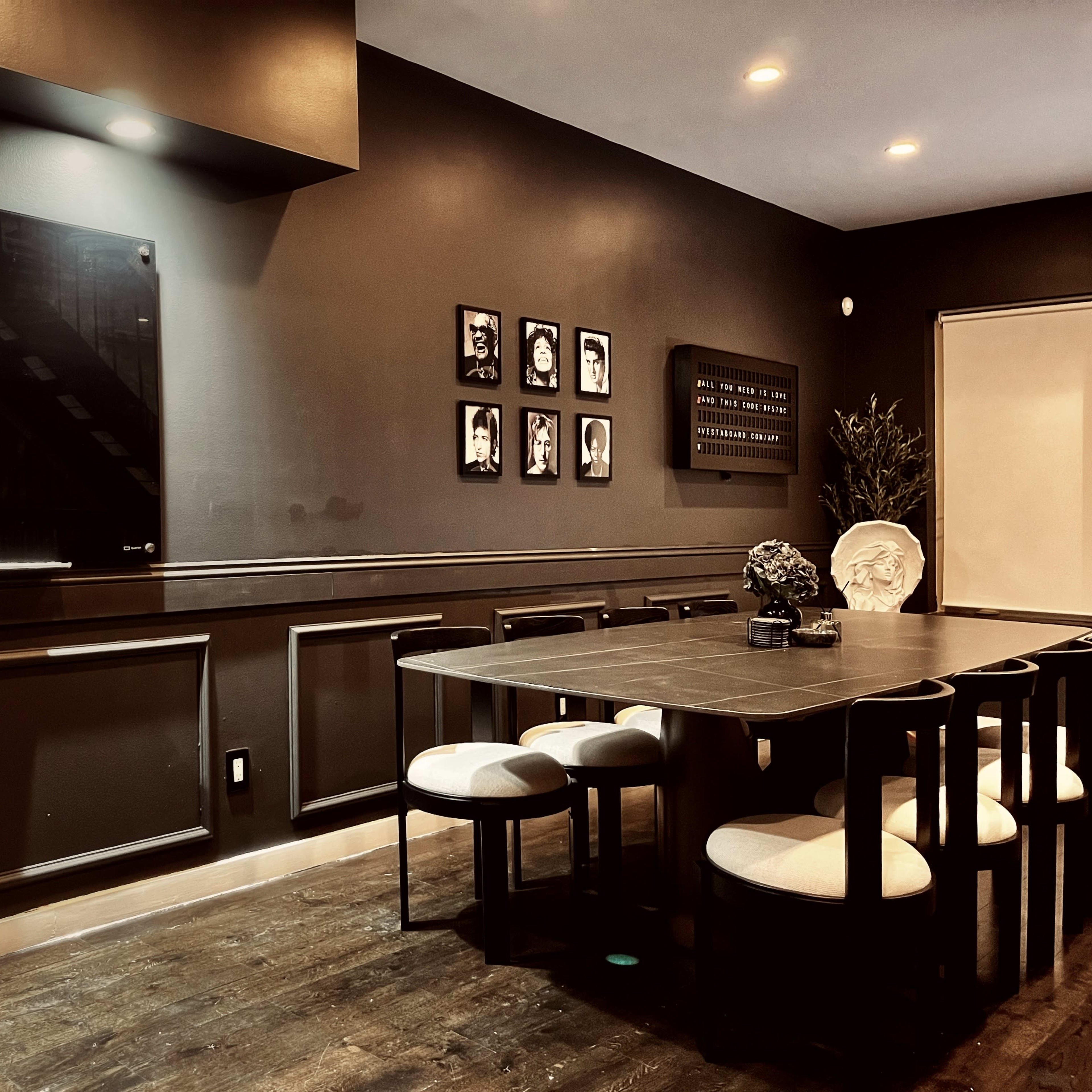 A dark-colored dining room features a long table surrounded by six chairs, with a television mounted on the wall and framed photographs displayed nearby.