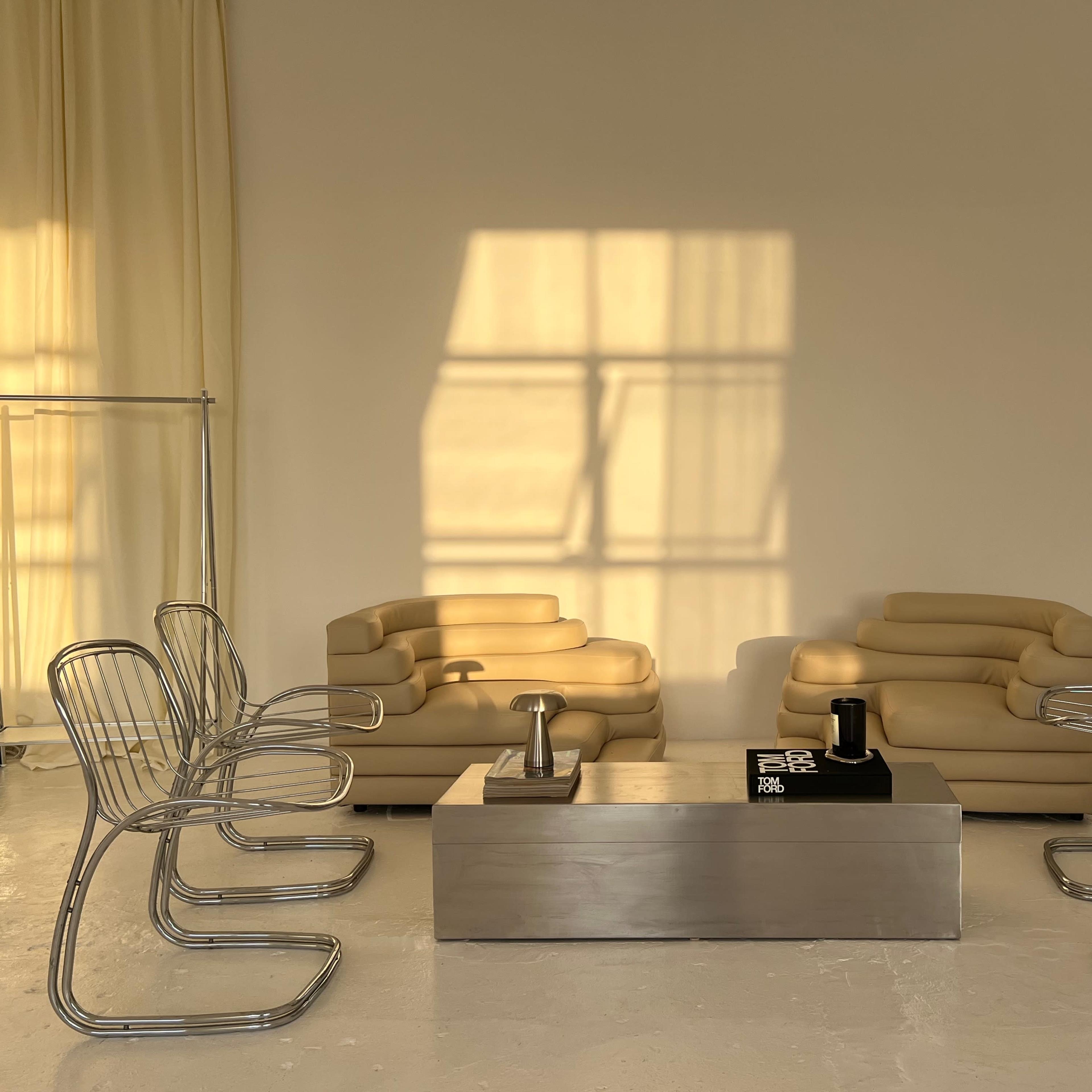 A minimalist seating area with two pale sofas facing each other, surrounded by six metallic chairs, and a rectangular coffee table in the center.
