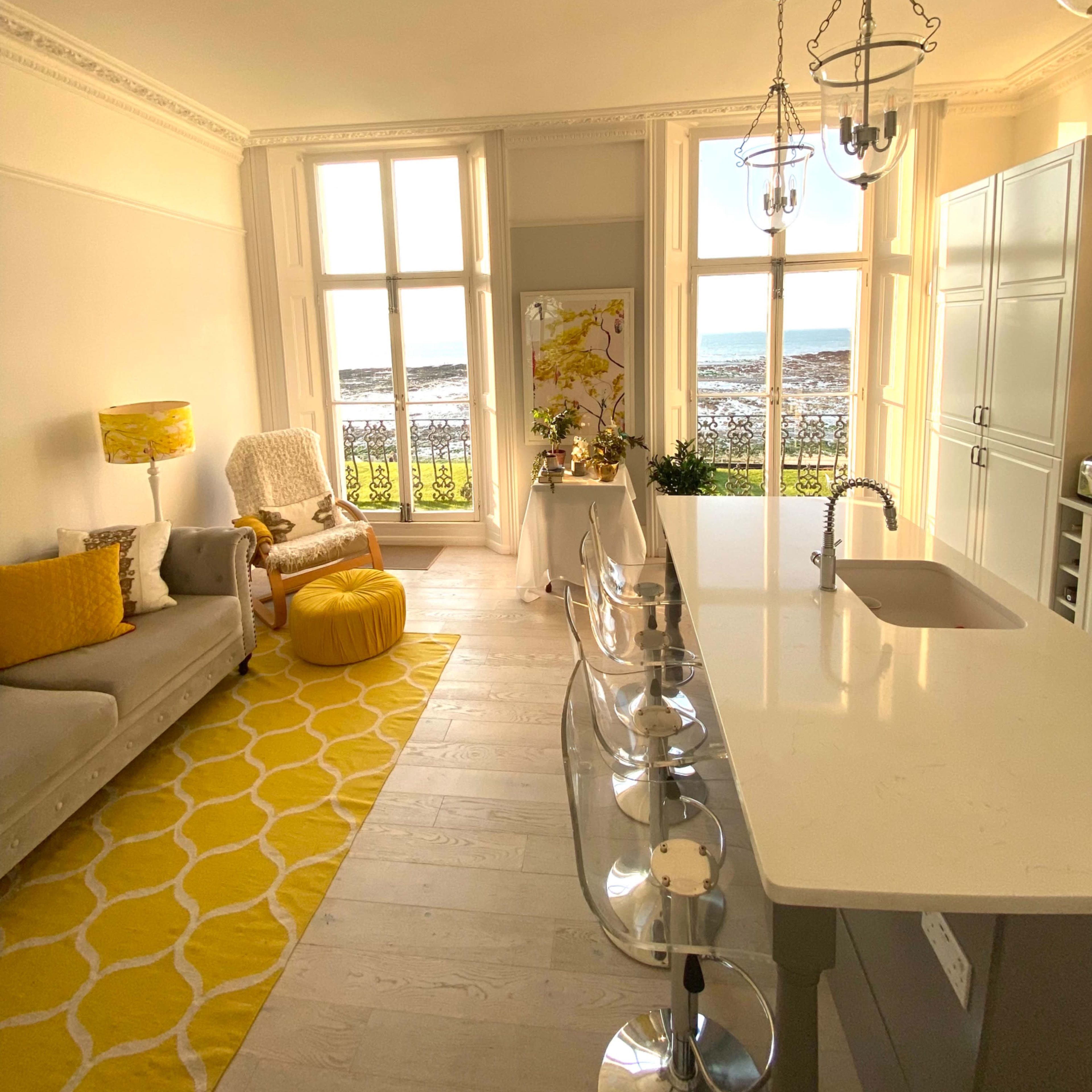 A bright kitchen and living area with large windows overlooking a body of water, featuring modern furnishings and a yellow color scheme.