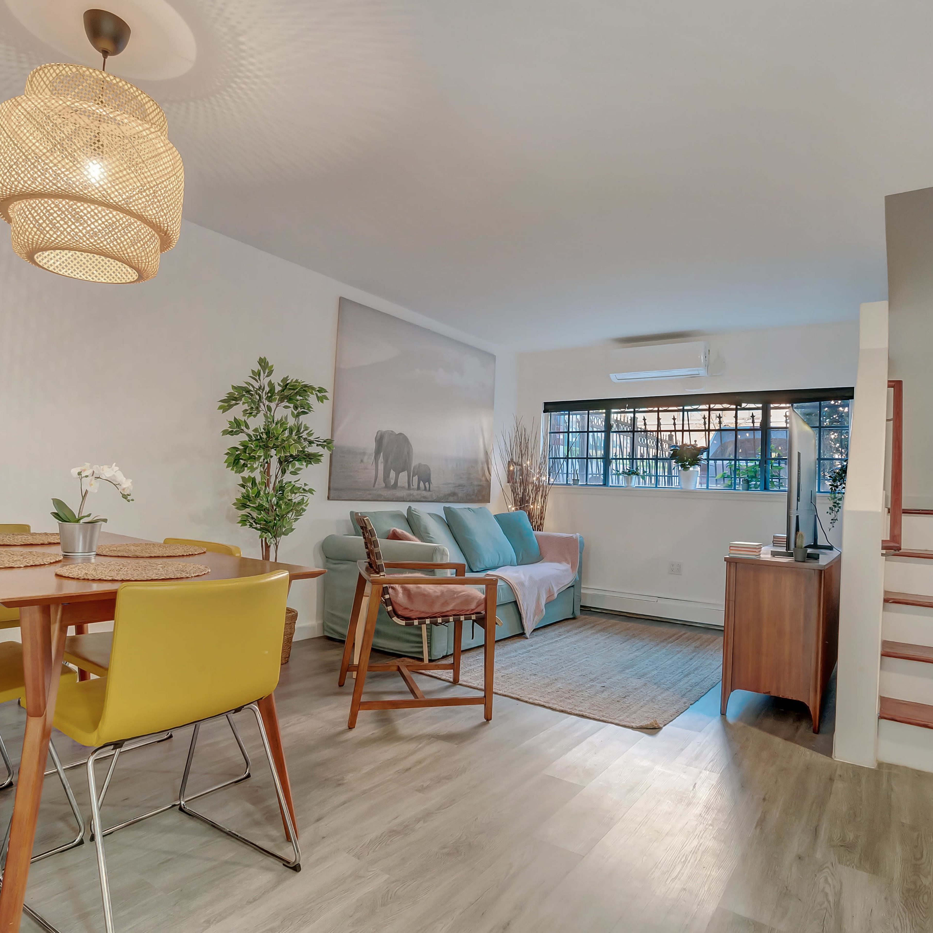 A bright living space featuring a dining area with yellow chairs, a sofa, a decorative plant, and a staircase leading to an upper level.