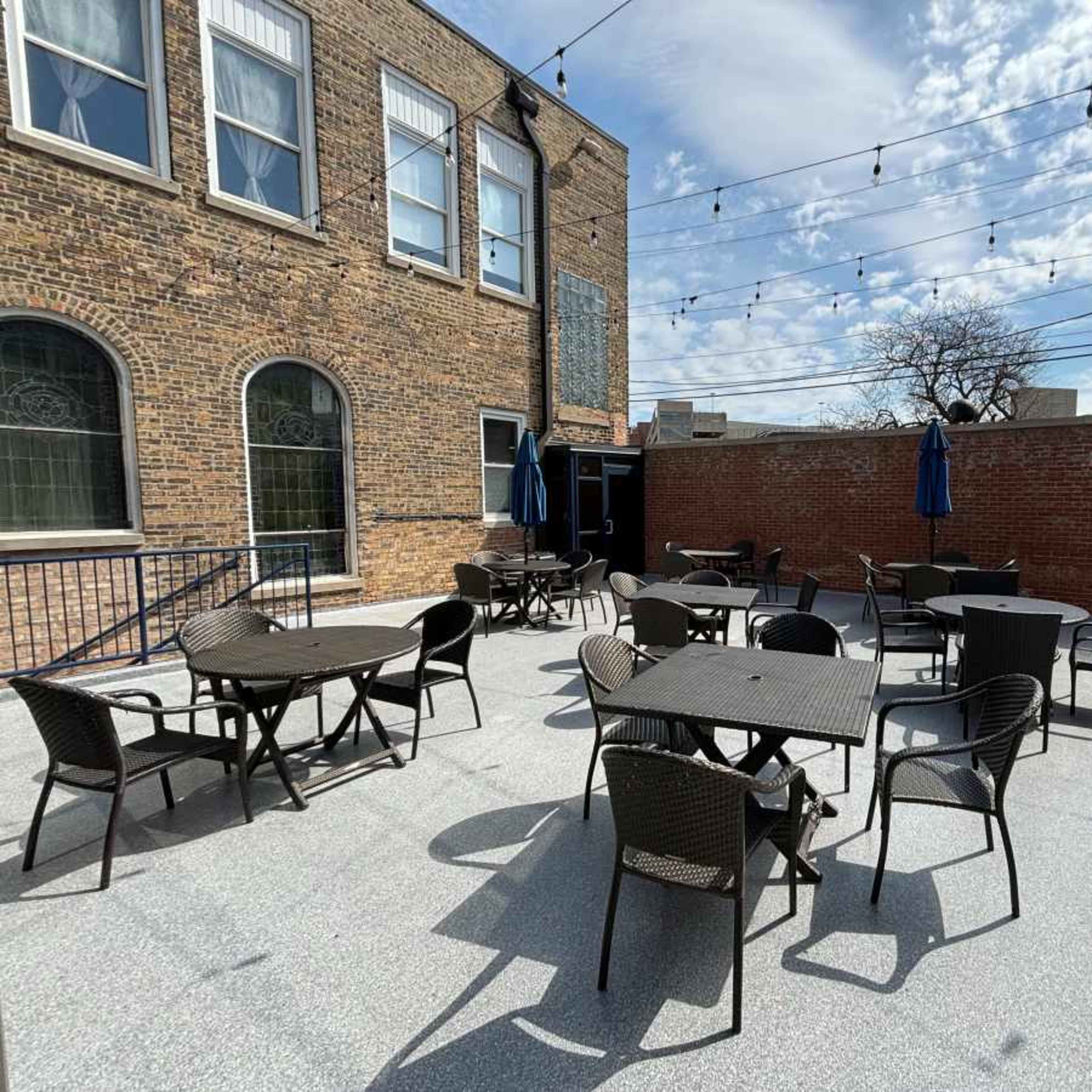 A rooftop patio with several dark tables and chairs arranged on a paved surface, surrounded by brick walls and string lights overhead.