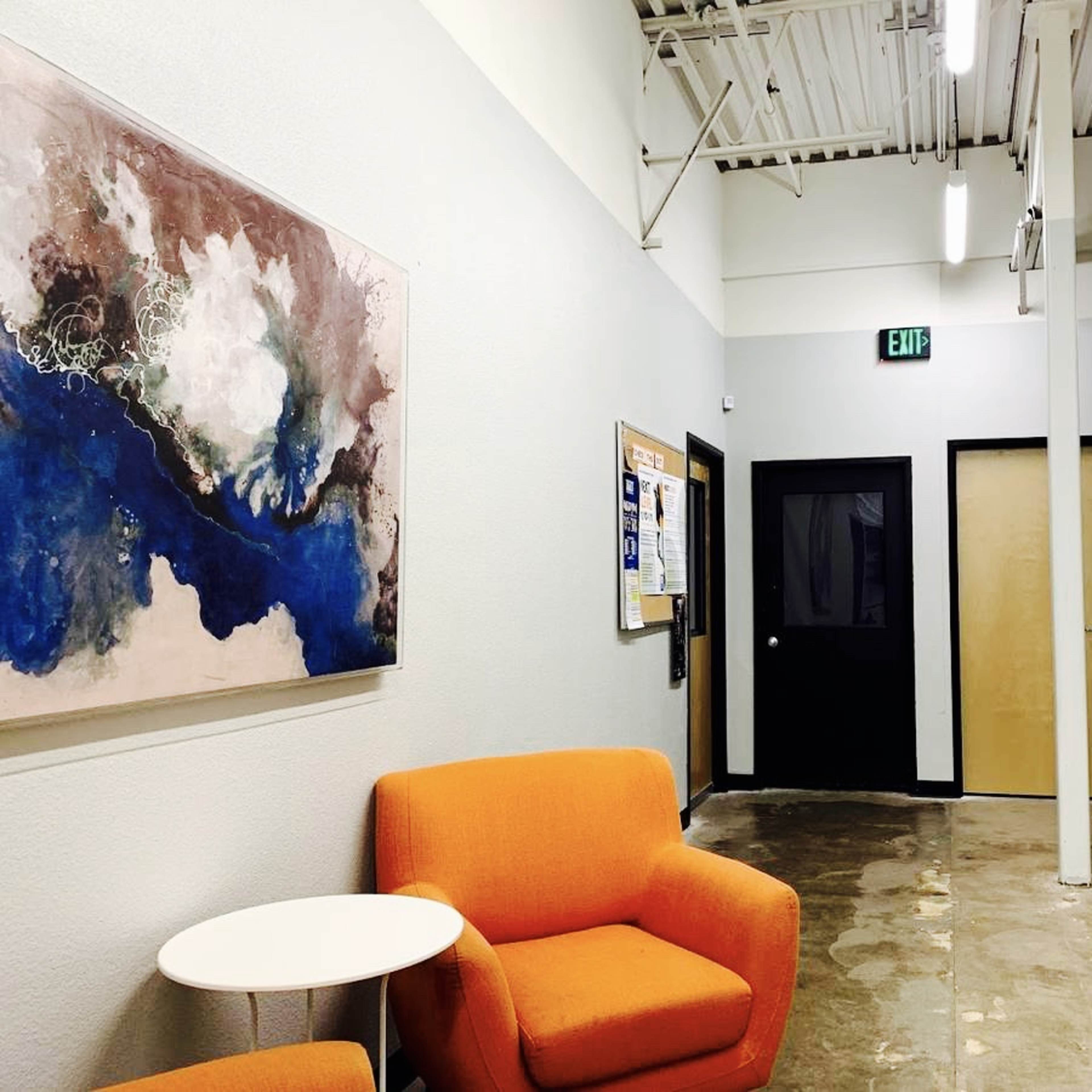 A modern hallway with two orange chairs and a small table, leading to doors and a bulletin board on the left wall.