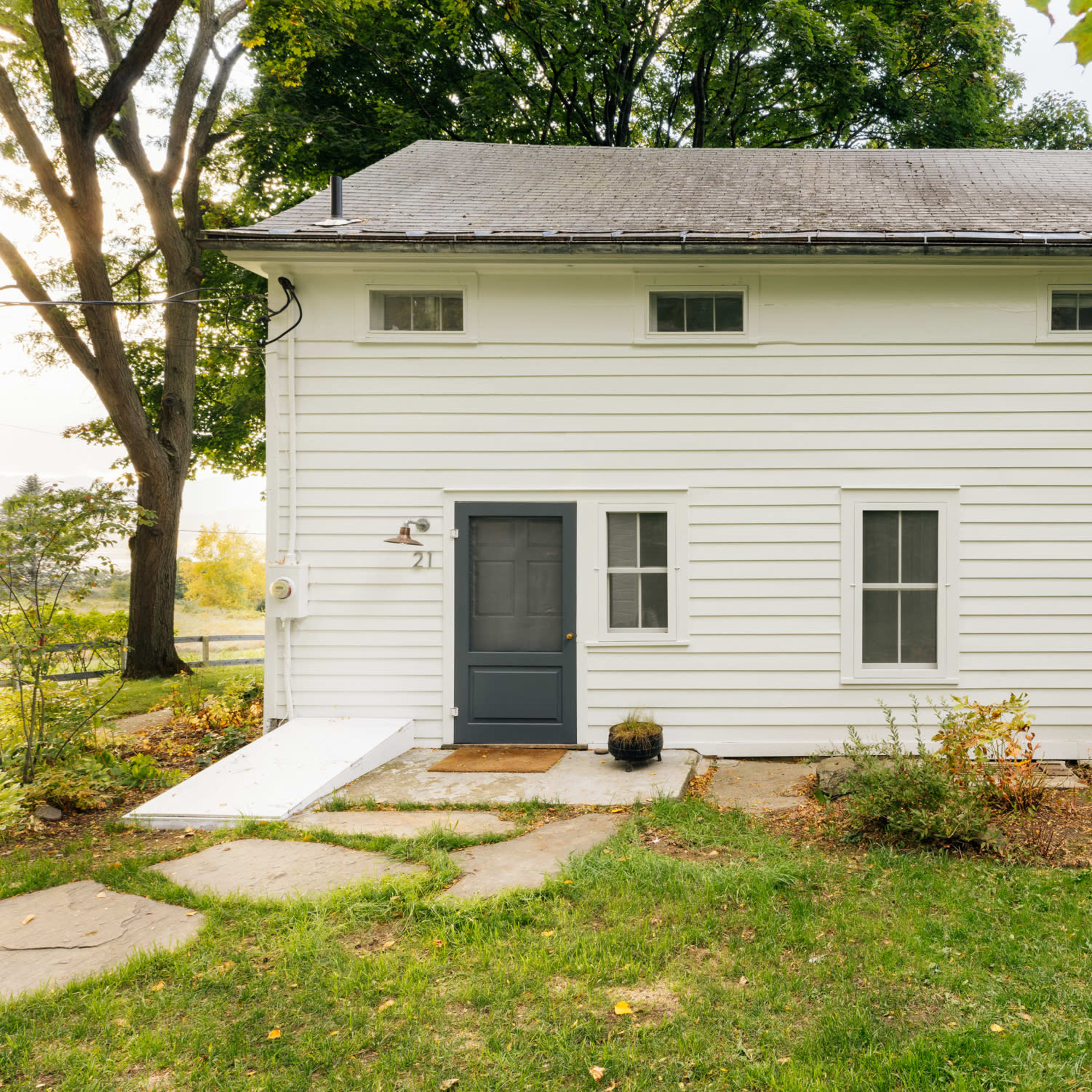 A white, two-story house with a dark gray front door and several small windows, set in a grassy area with a concrete path leading up to it.