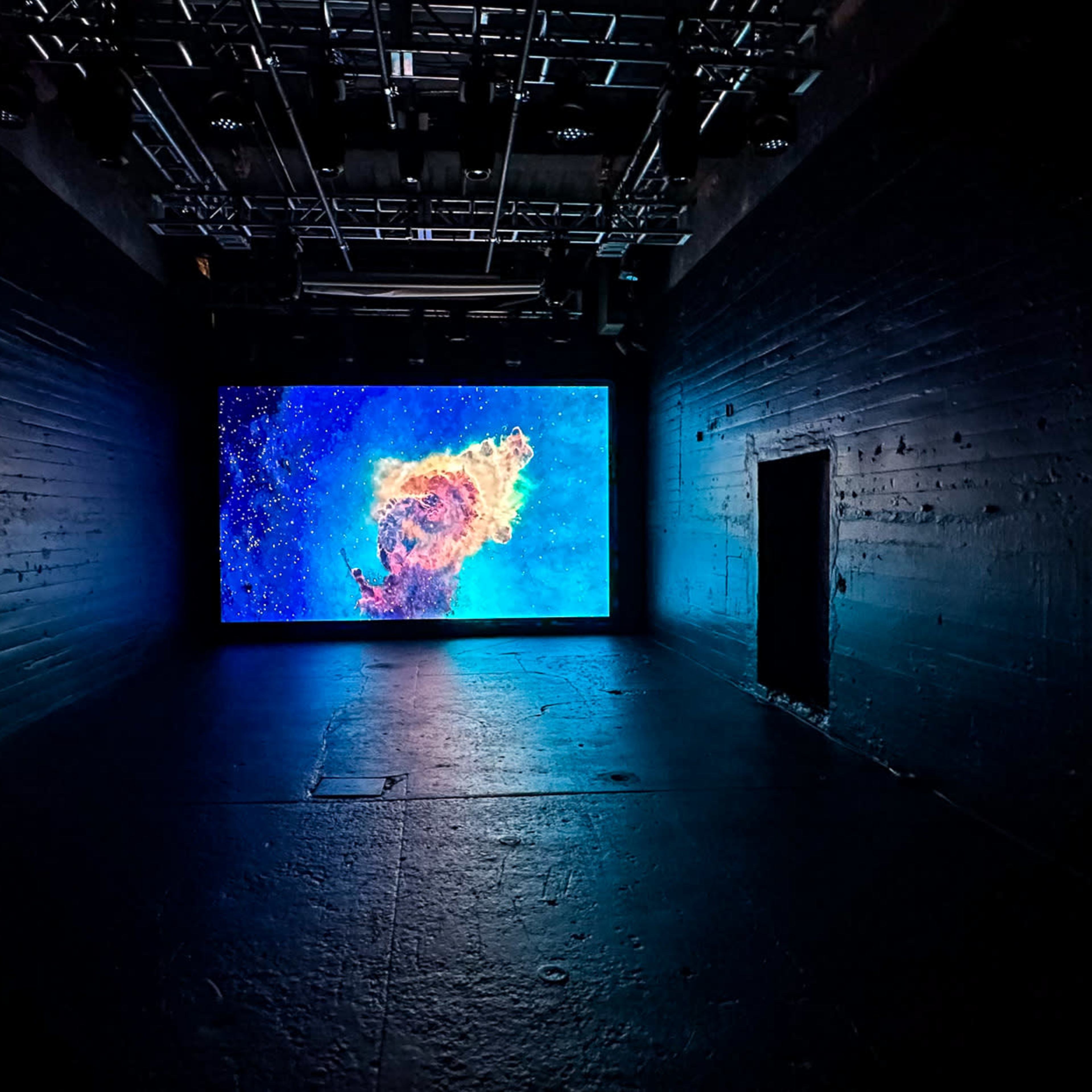 A large screen displaying colorful visuals is positioned in a dark, industrial-looking room with textured walls.