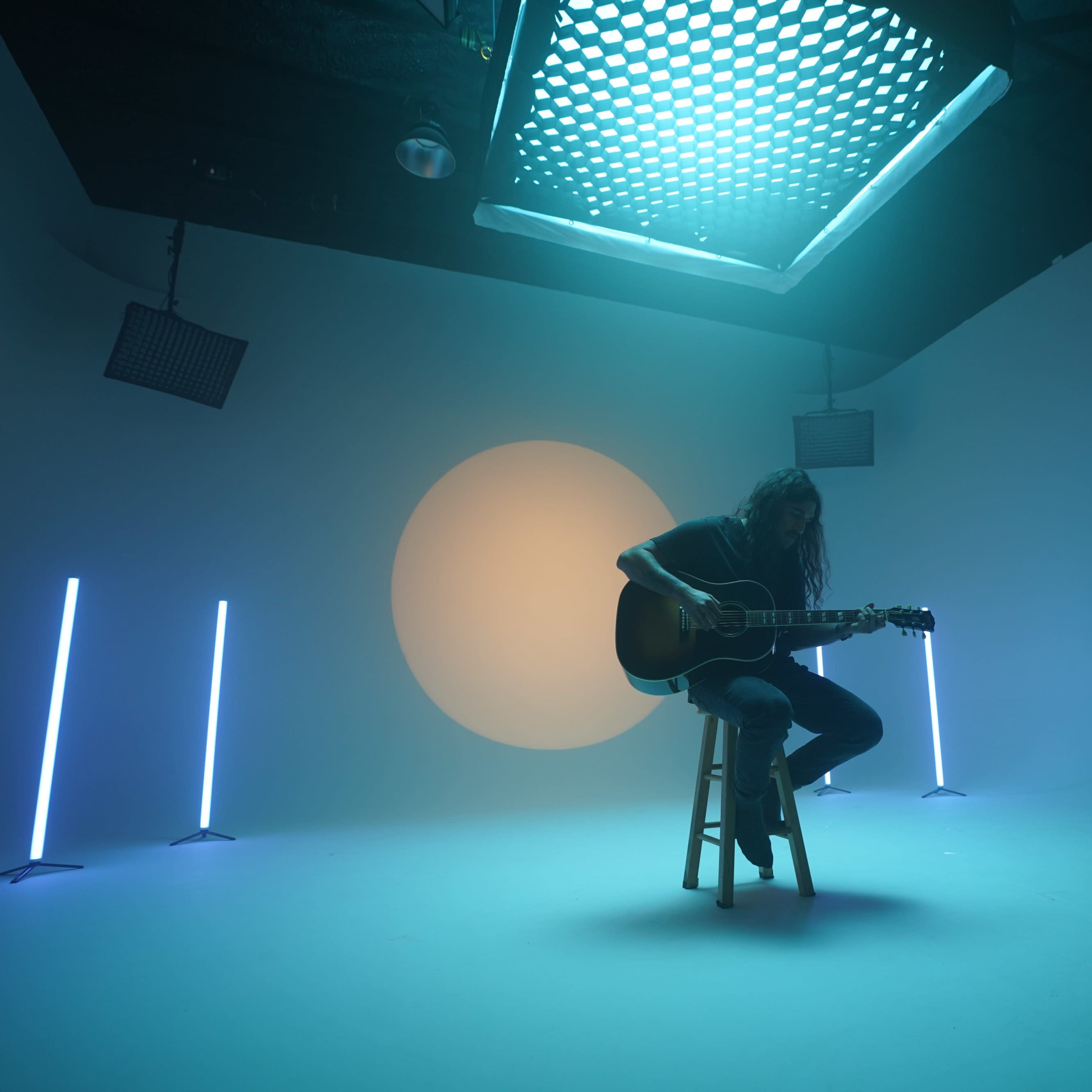 A musician plays an acoustic guitar while seated on a stool in a dimly lit studio with colored lighting and a large circular backdrop.