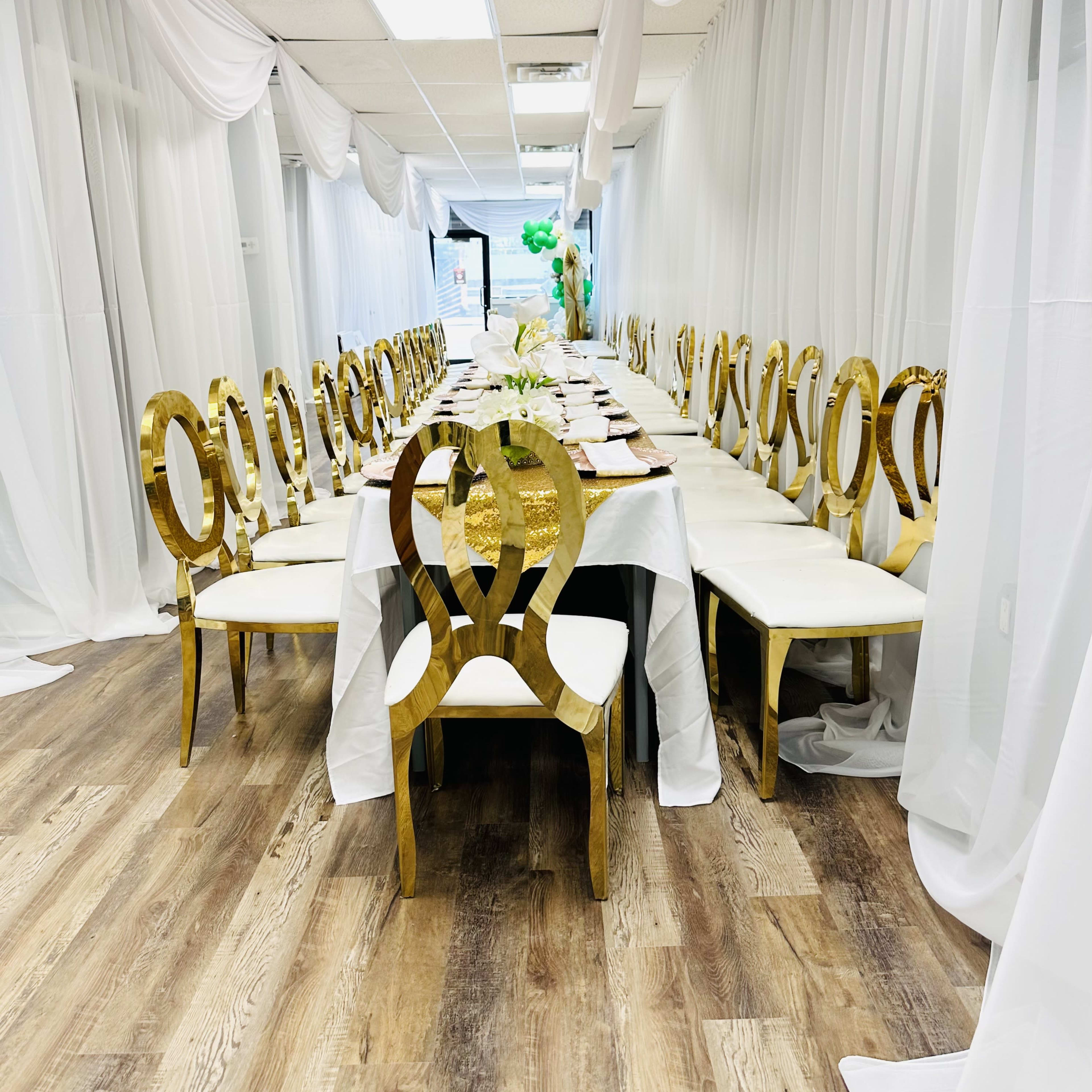 A long dining table with gold chairs is set up in a hall adorned with white drapery.