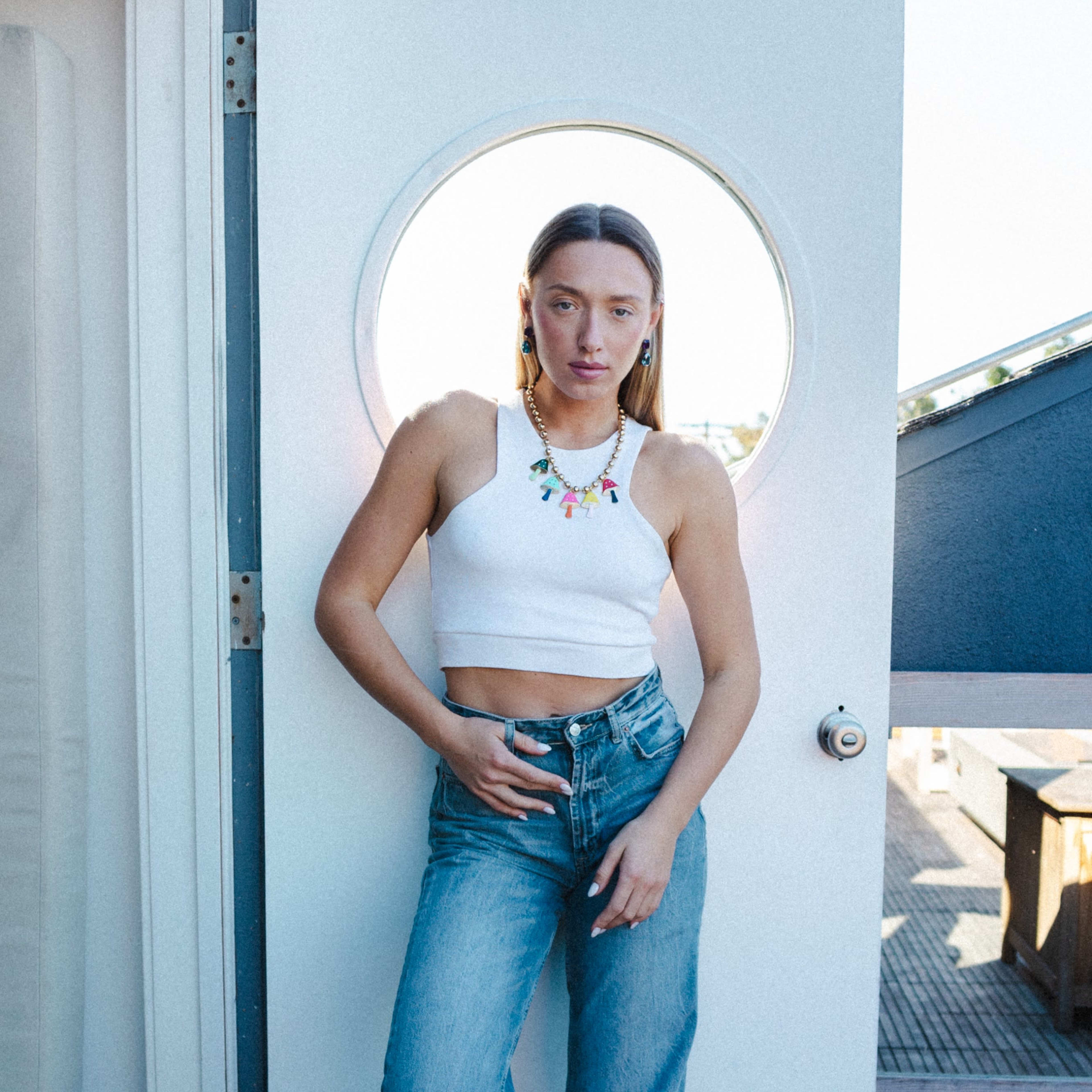 A person stands in front of a circular doorway, wearing a white crop top and blue jeans, with a colorful necklace.
