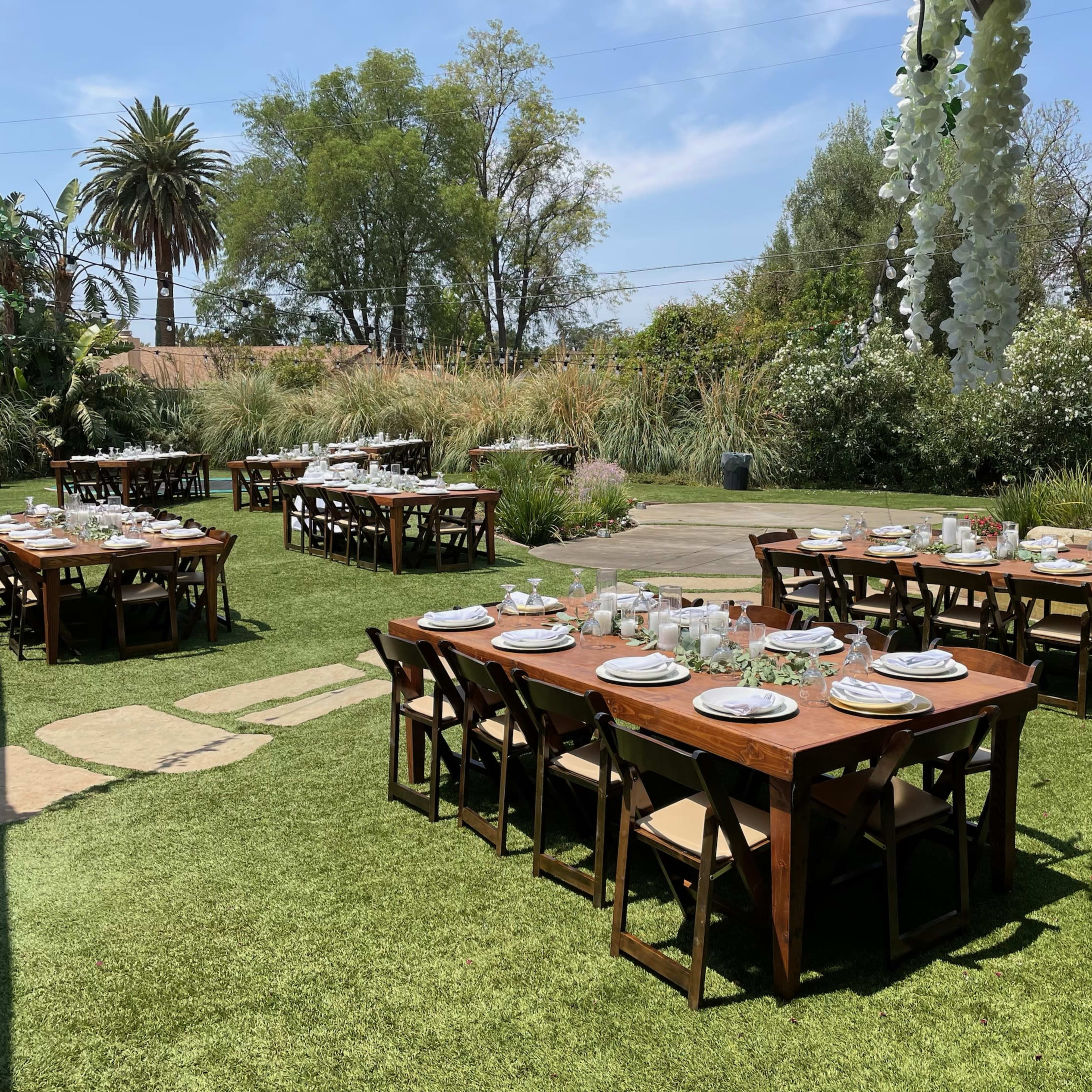 A setup for an outdoor dining event with multiple wooden tables arranged on a grass lawn, surrounded by greenery and palm trees.